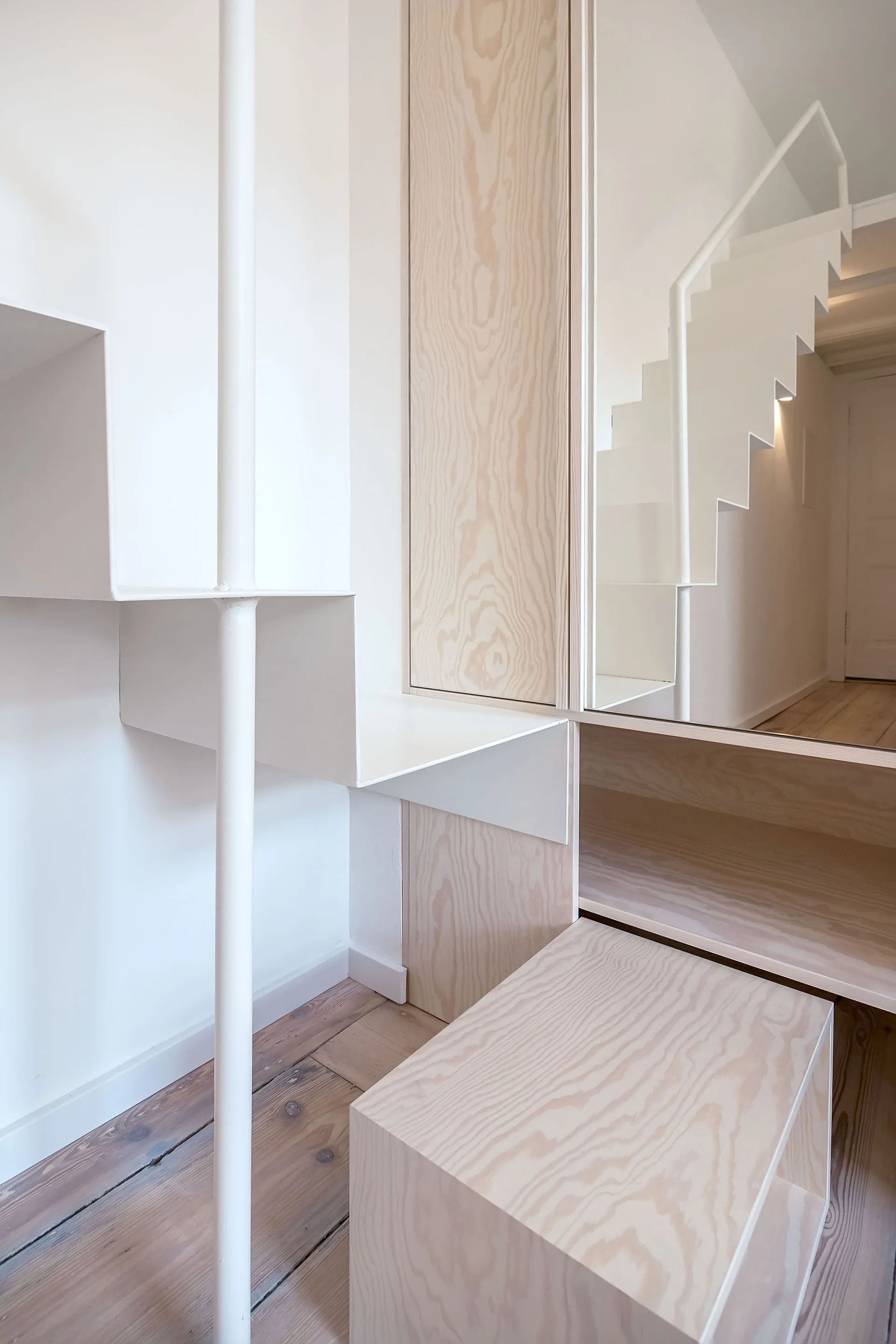 A minimalist white metal staircase features integrated natural wood storage cabinets, a wall mirror, and light wood flooring.
