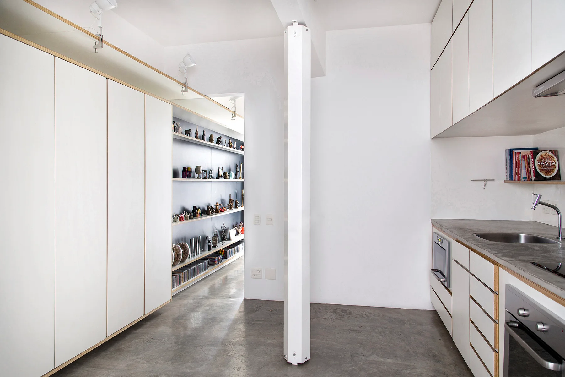 A minimalist kitchen features white laminate cabinetry with exposed plywood edges, a gray concrete floor, and a central white support column.