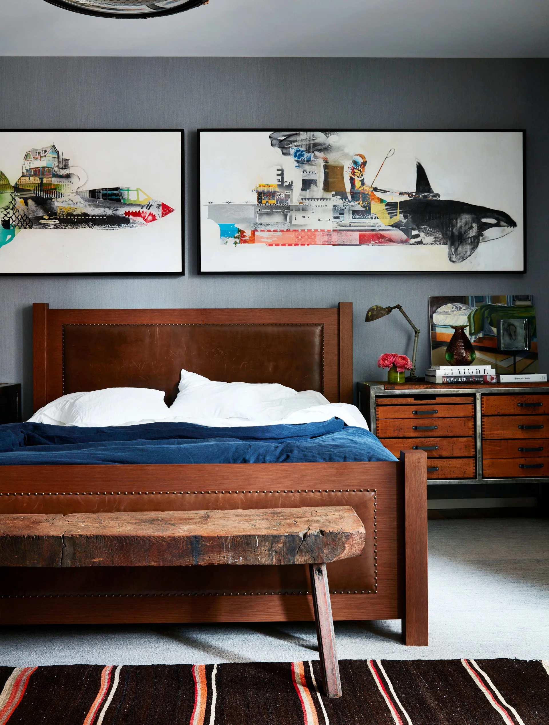 A bedroom features a brown leather headboard, a rustic wooden bench, and two large framed art prints hanging on a dark grey wall.