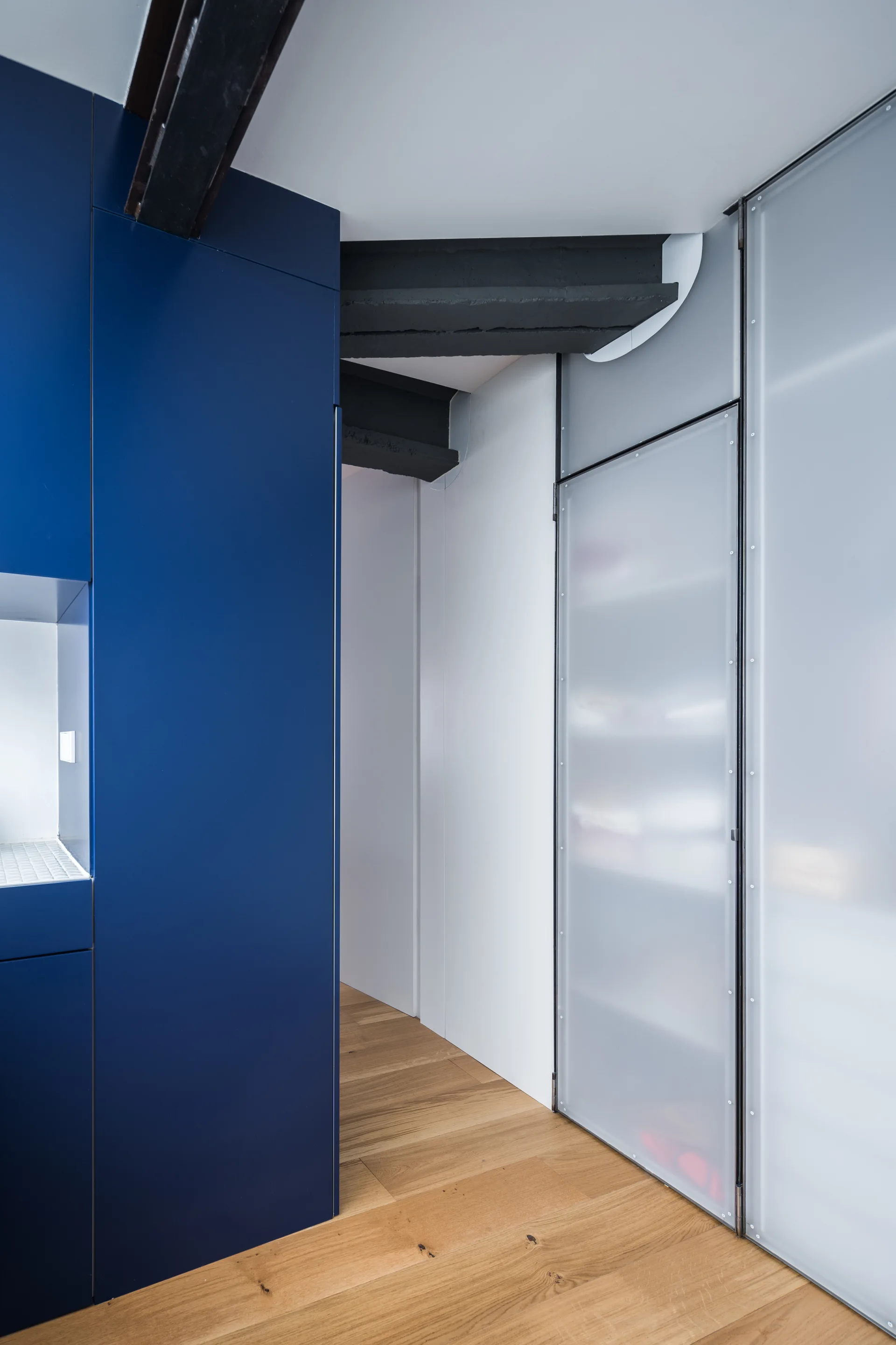 A hallway features matte navy blue cabinetry, a frosted glass storage door, and exposed black steel ceiling beams over light oak wood floors.