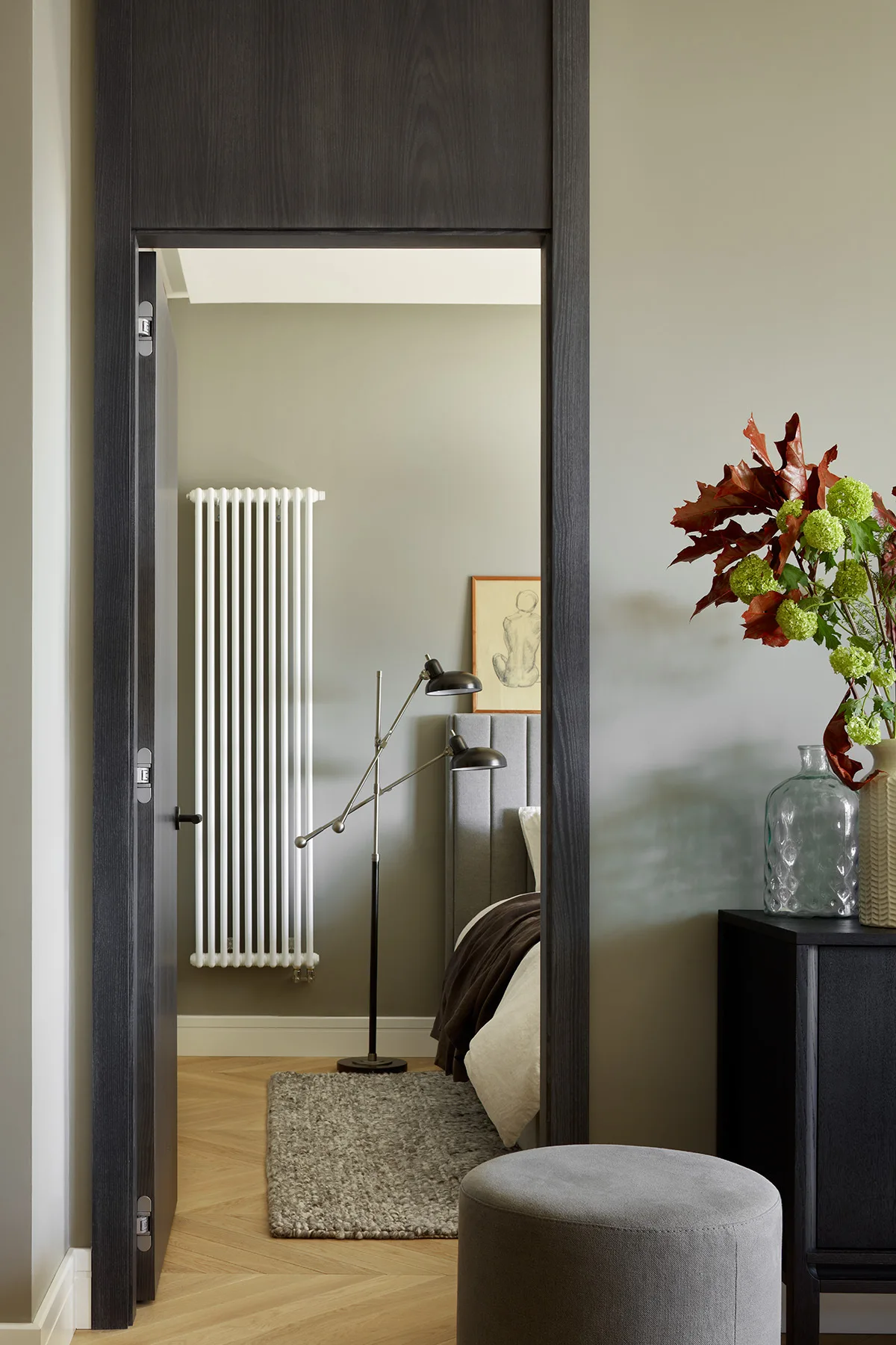 A view through a dark charcoal doorway into a sage green bedroom with a white vertical radiator and herringbone hardwood floors.