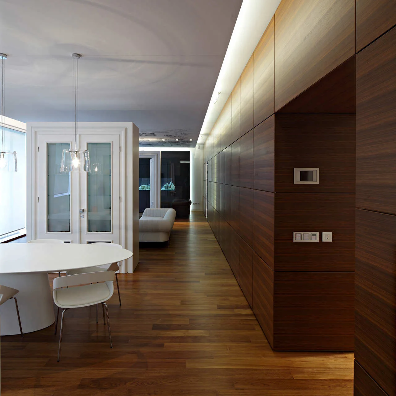 A white oval dining table and modern chairs stand next to a long wall of dark wood paneling illuminated by warm cove lighting.