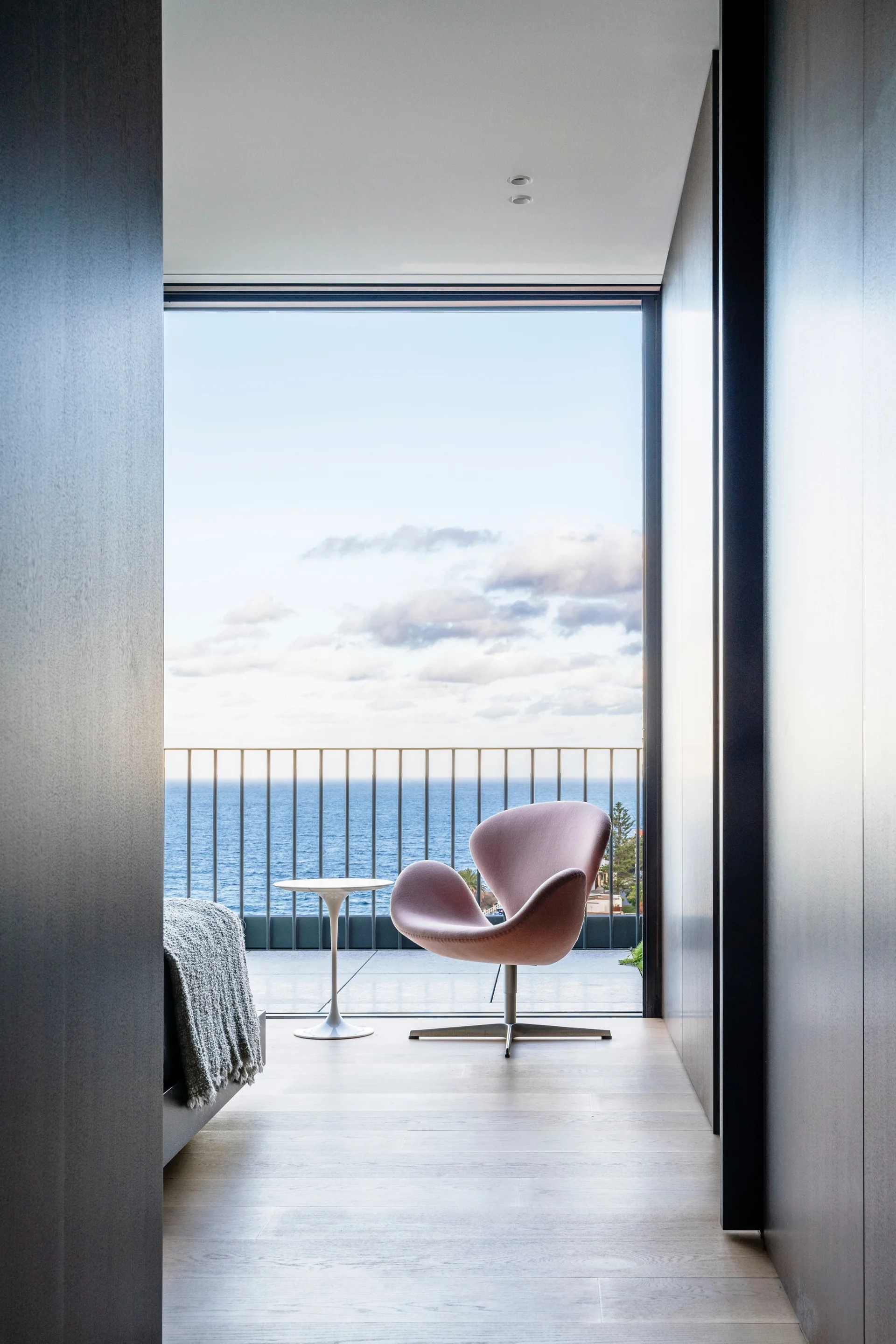 A pink swan chair and small white side table sit by a large window overlooking the ocean from a bedroom with light wood floors.