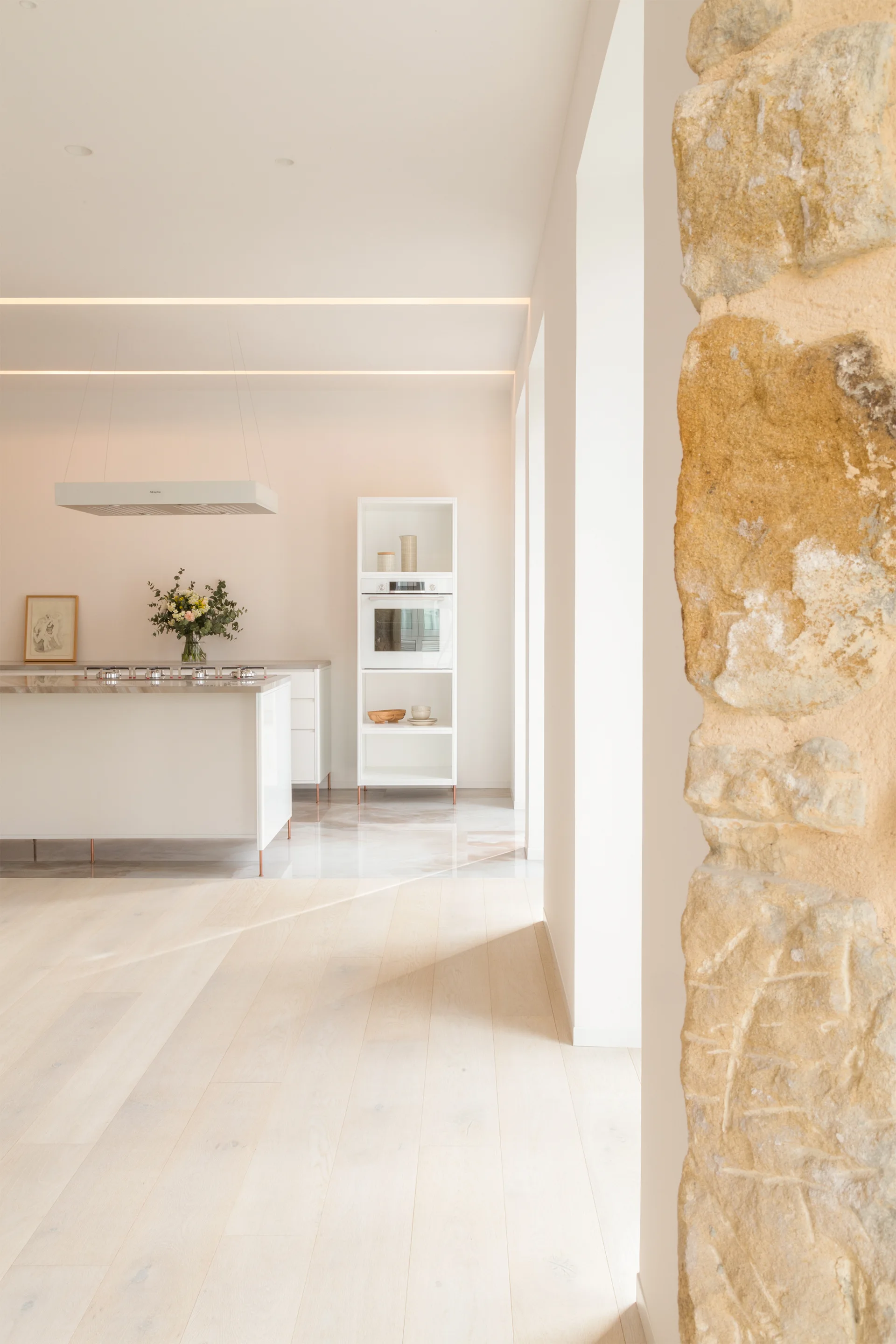 A minimalist white kitchen featuring a central island, integrated appliances, blonde wood floors, and a textured natural stone column in the foreground.