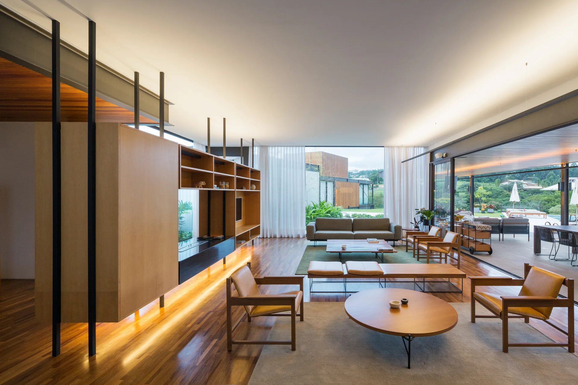 A spacious living room with warm wood floors, a floating partition with integrated shelving, leather armchairs, and floor-to-ceiling glass walls.