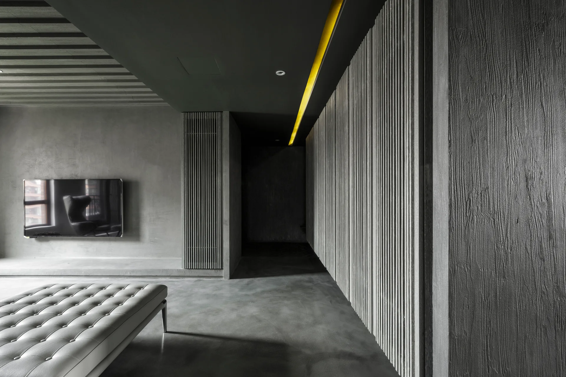 A minimalist gray lounge featuring a tufted bench, smooth concrete walls, a dark wood slat wall, and a yellow recessed ceiling light strip.