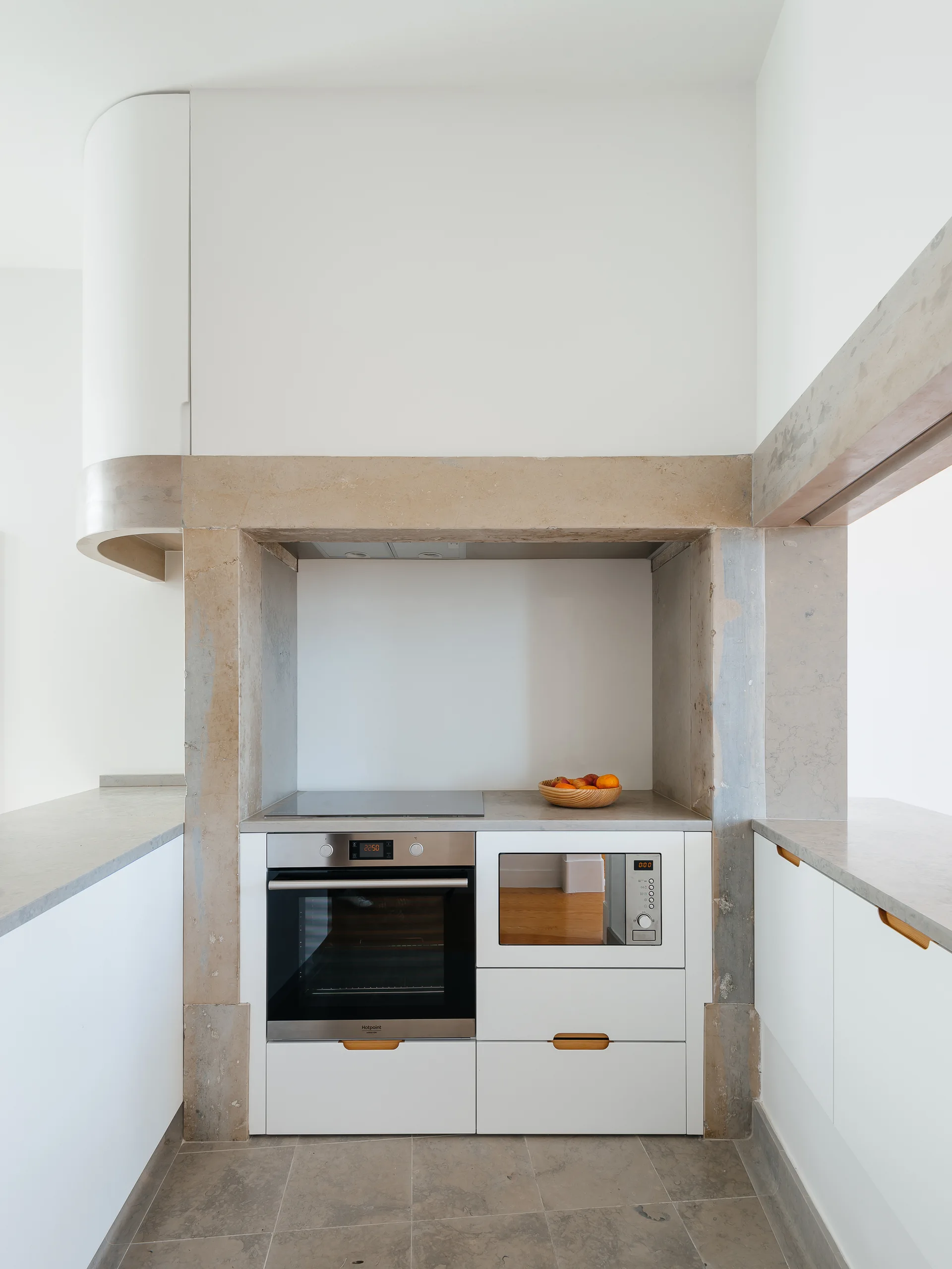 A modern kitchen featuring white flat-panel cabinets and appliances integrated into a large, rectangular beige stone architectural frame.
