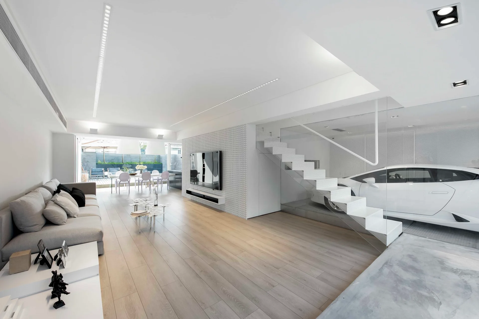 A minimalist white living room features a grey sectional sofa, a floating white staircase, and a sports car visible behind a glass partition.
