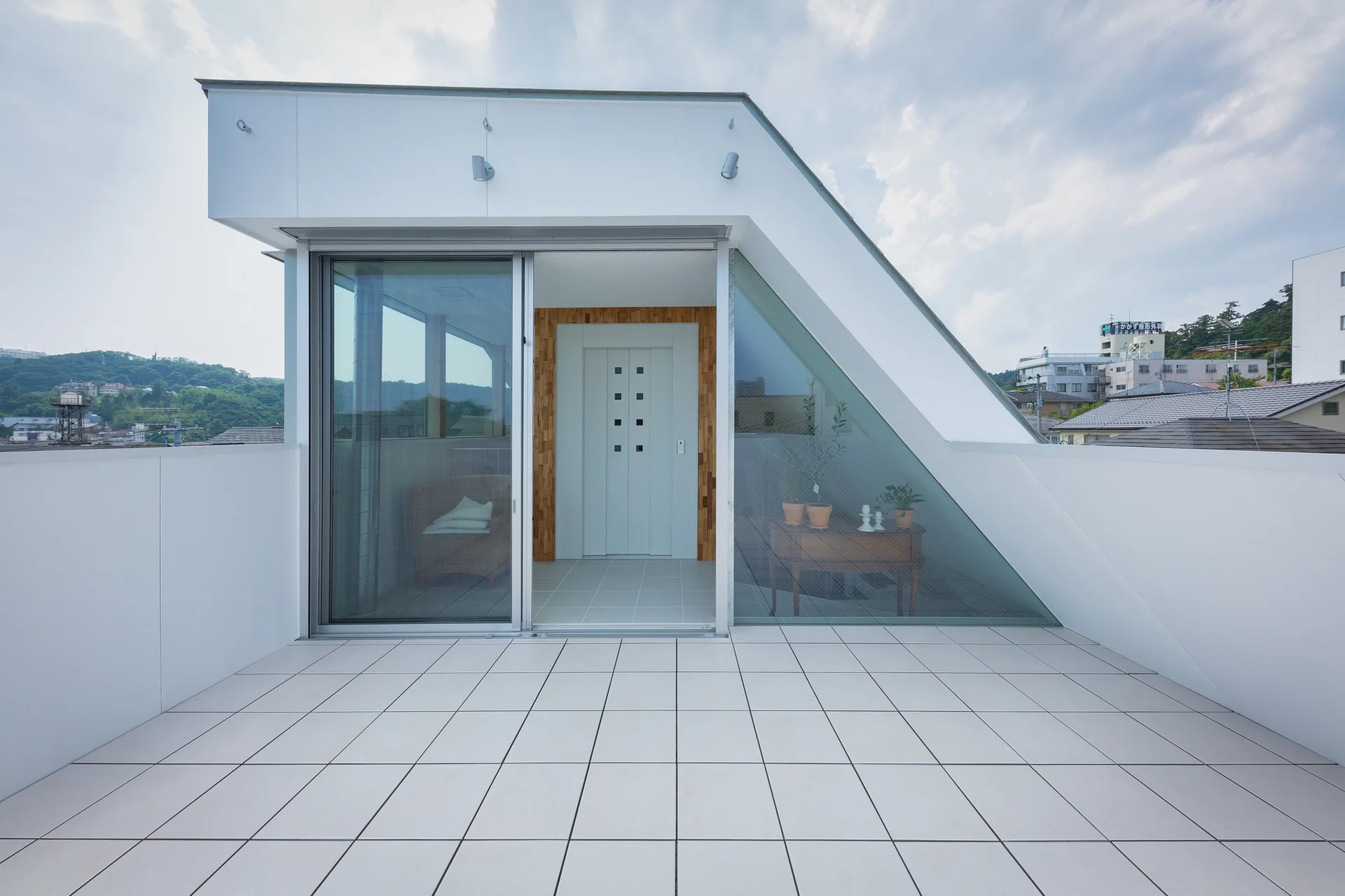 A modern outdoor terrace with white floor tiles, a slanted roofline, and sliding glass doors revealing an interior entryway.
