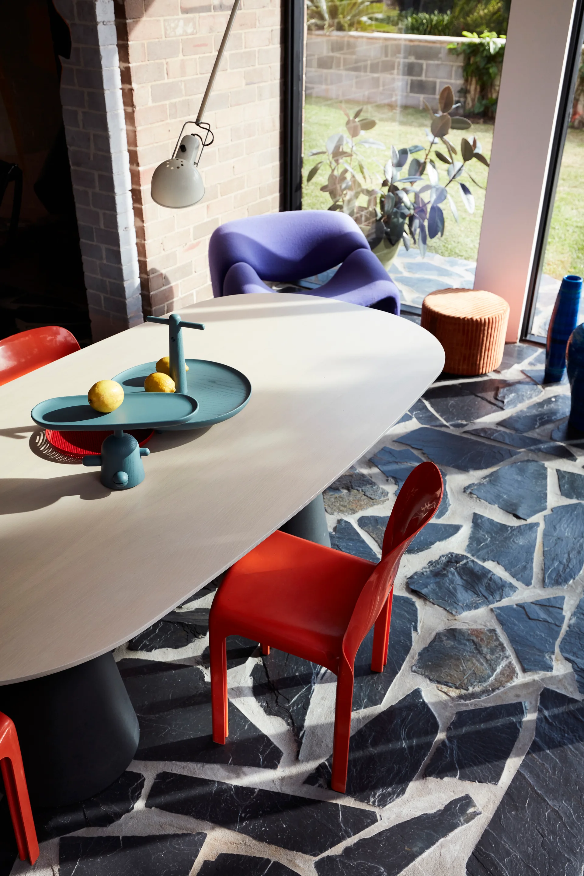 An oval light wood dining table with bright orange chairs sits on a dark flagstone floor next to floor-to-ceiling windows.