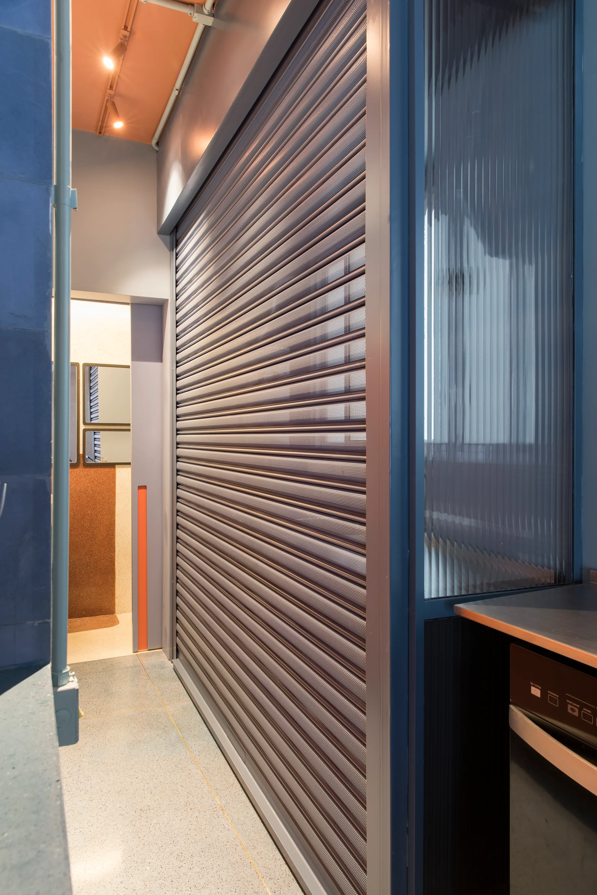 A modern corridor features a grey perforated metal rolling shutter door next to a fluted glass partition and a speckled terrazzo floor.