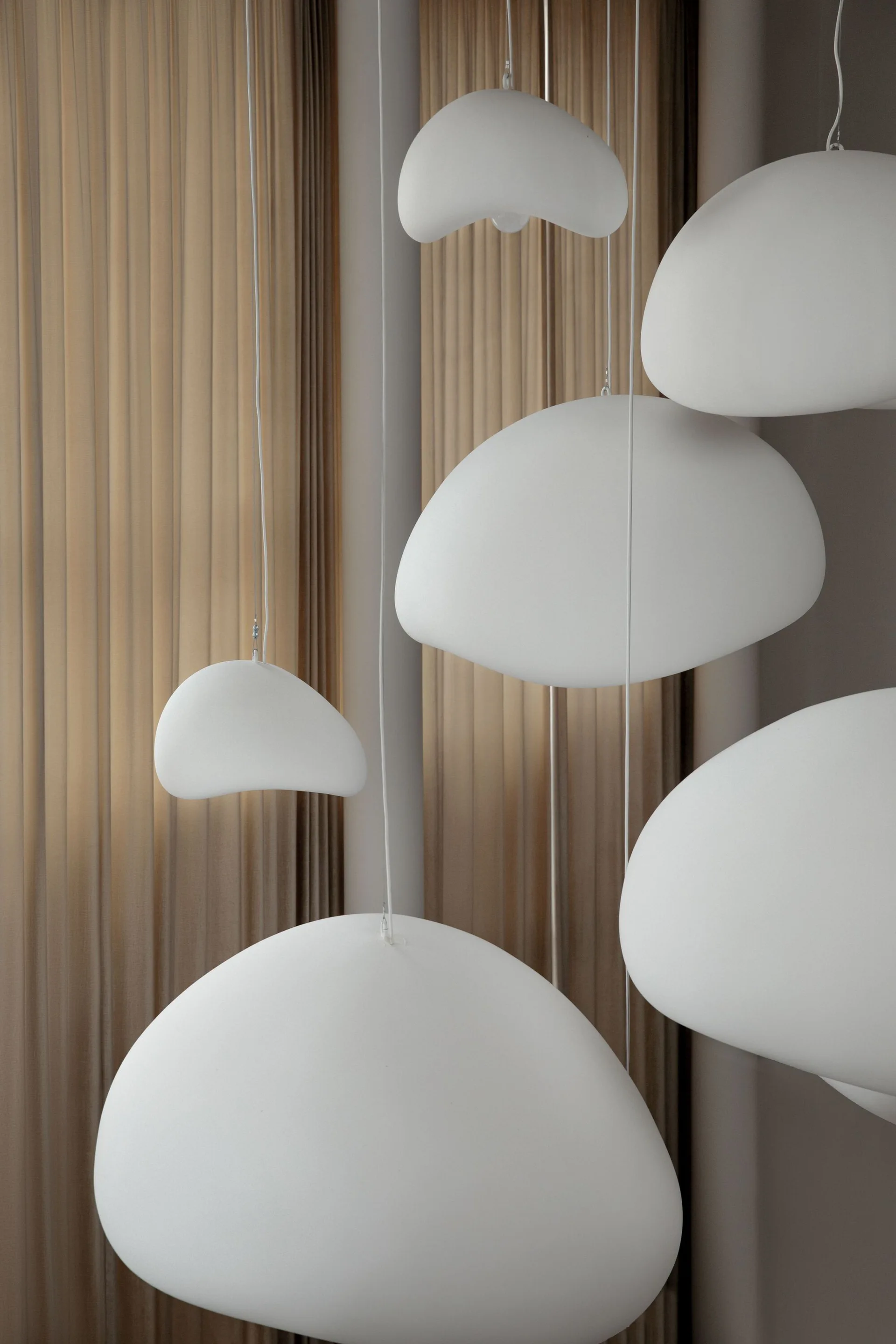 Several matte white pendant lights in organic pebble shapes hang at staggered heights in front of pleated beige curtains.