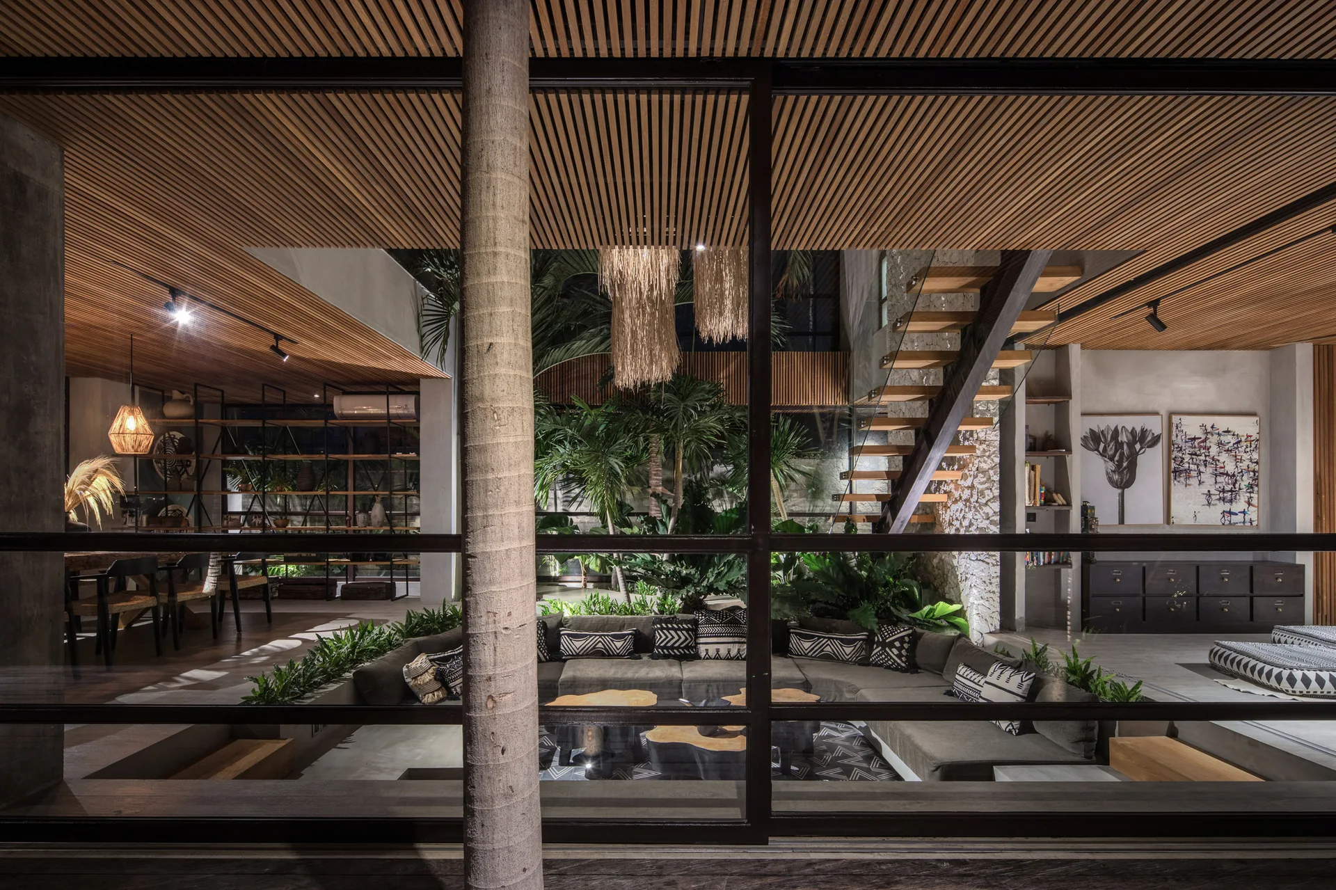 An interior view of a tropical living room with a slatted wood ceiling, a u-shaped sectional sofa, an indoor garden, and a floating wooden staircase.