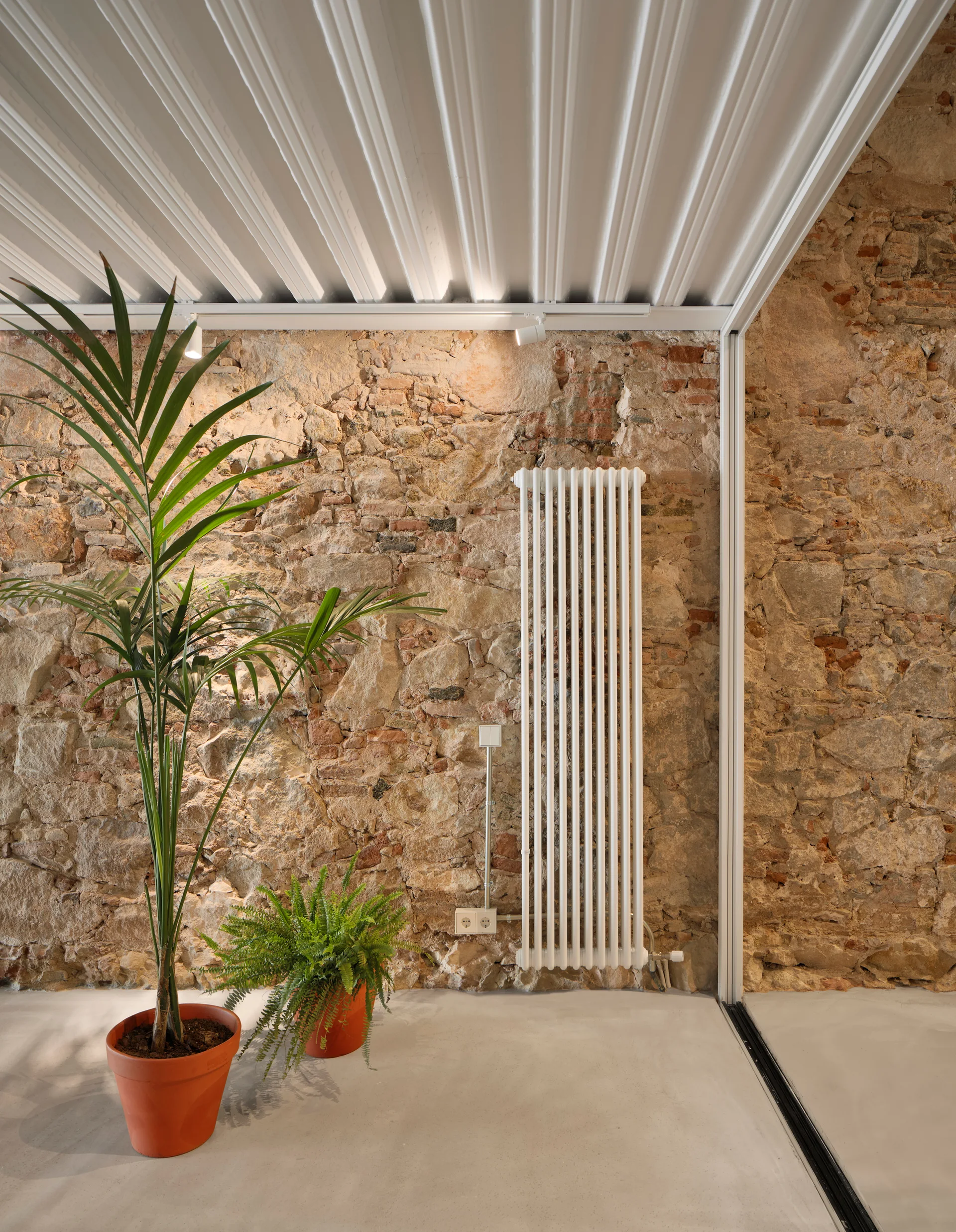 A white column radiator is mounted on a rough exposed stone wall next to terracotta potted plants under a white corrugated ceiling.