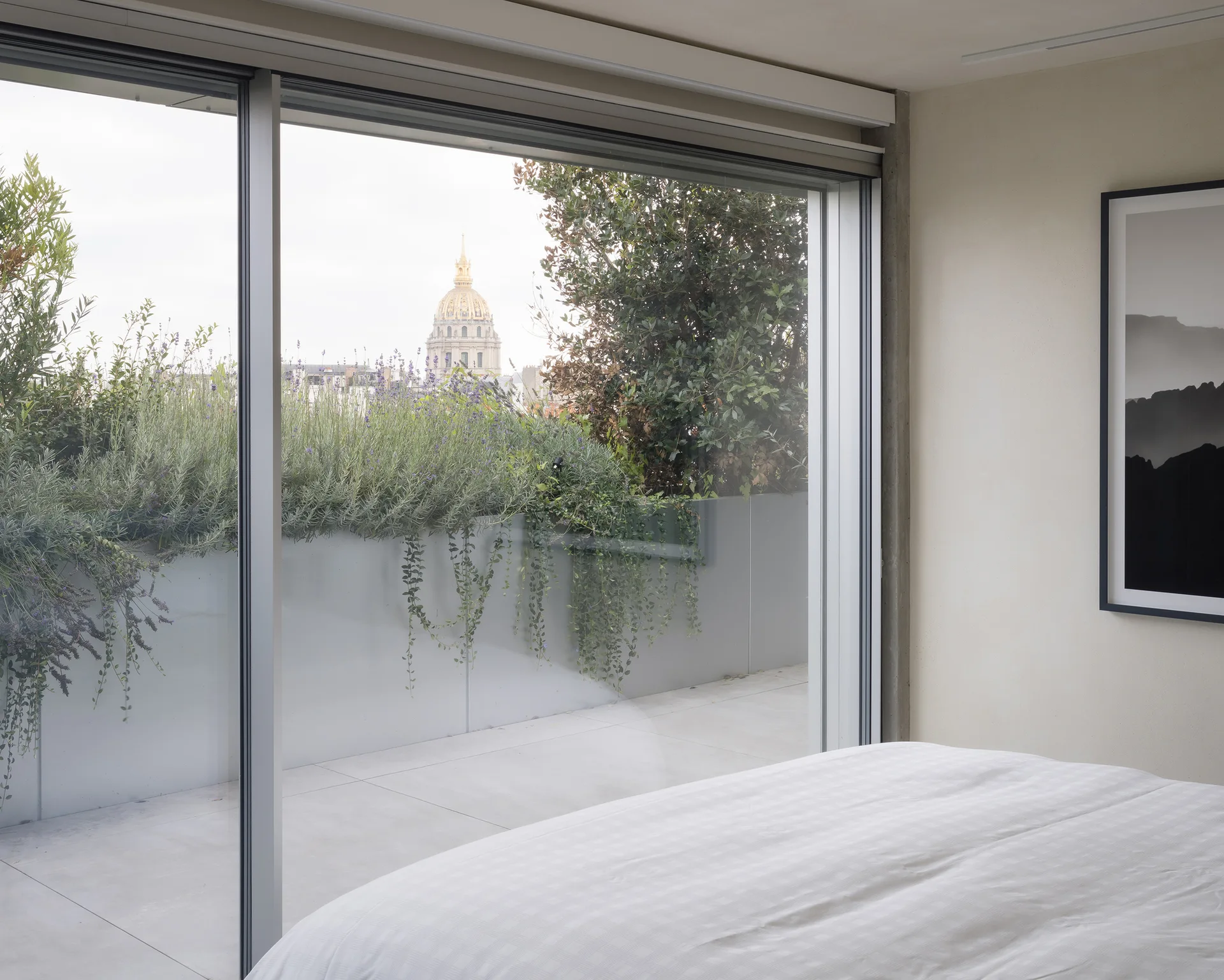 A minimalist bedroom featuring white grid-patterned bedding next to a large sliding glass door that opens to a terrace with green planters.