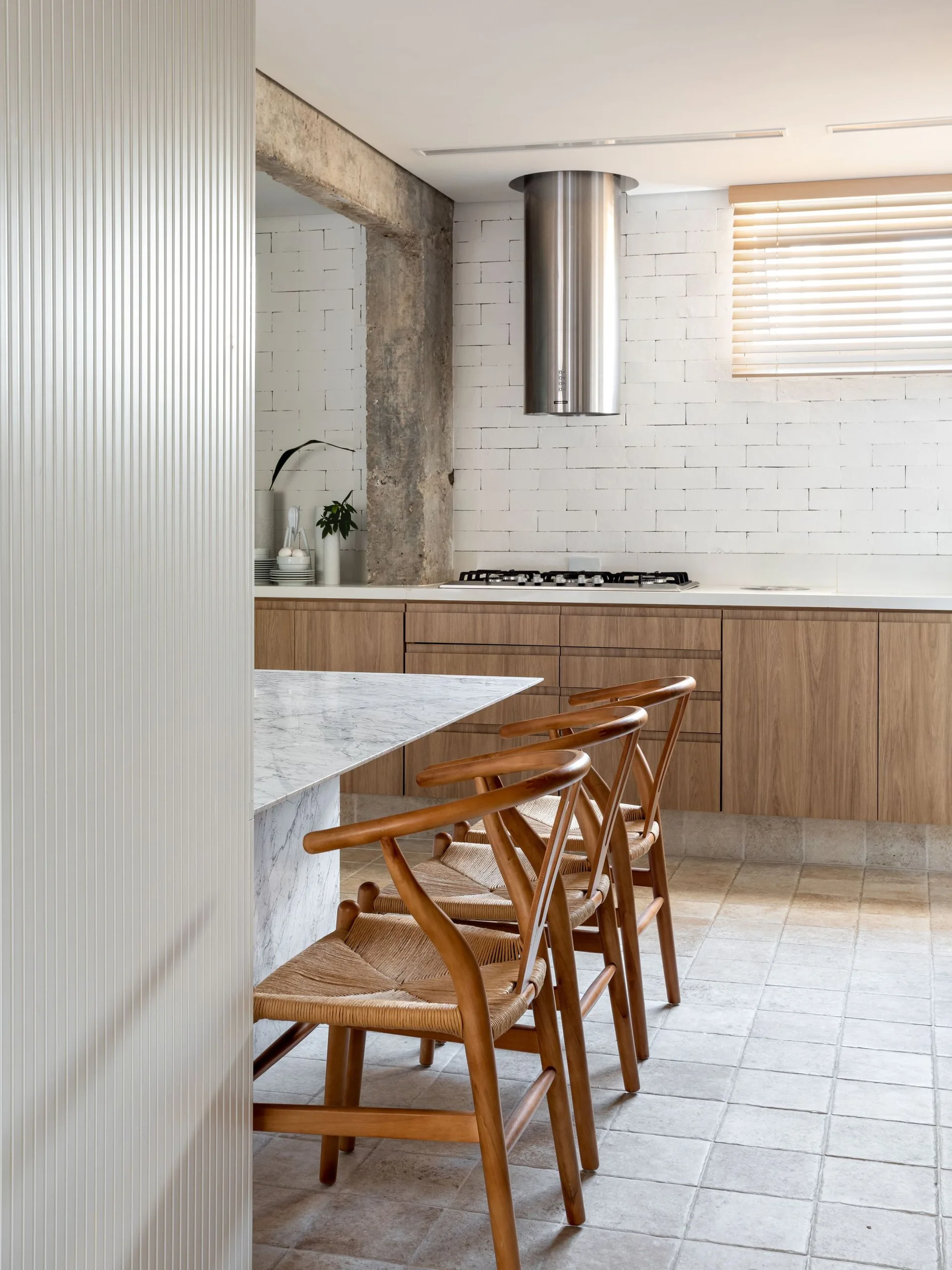 Three wooden wishbone stools sit at a marble island in a kitchen featuring light wood cabinets, a white brick wall, and an exposed concrete column.