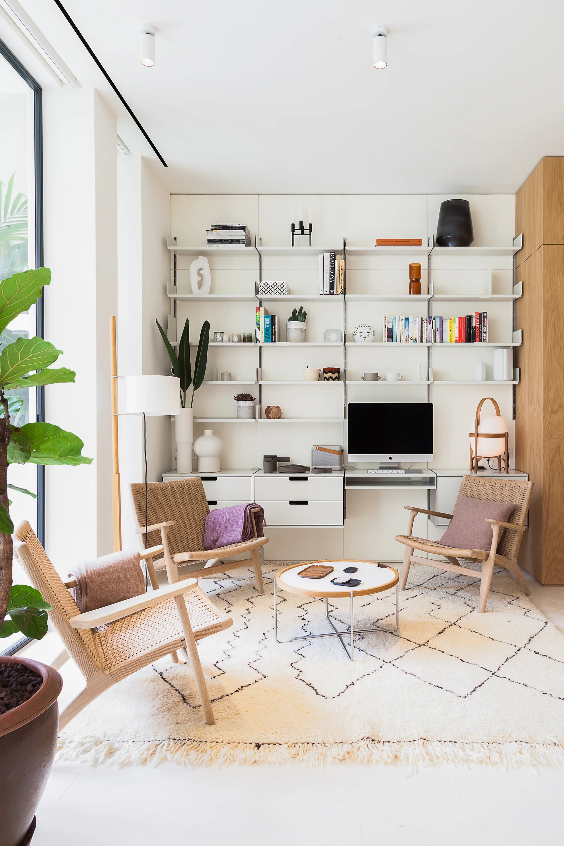 A bright living room features a white modular shelving unit with an integrated desk, three woven wooden armchairs, and a cream diamond pattern shag rug.