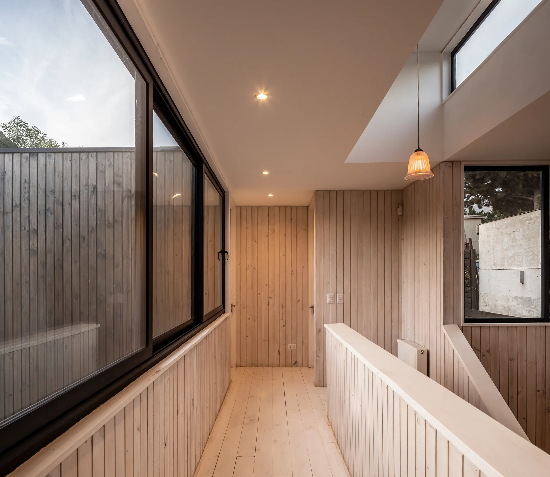 A minimalist hallway lined with vertical pale wood paneling features a black-framed window wall, wood plank floors, and a staircase railing.