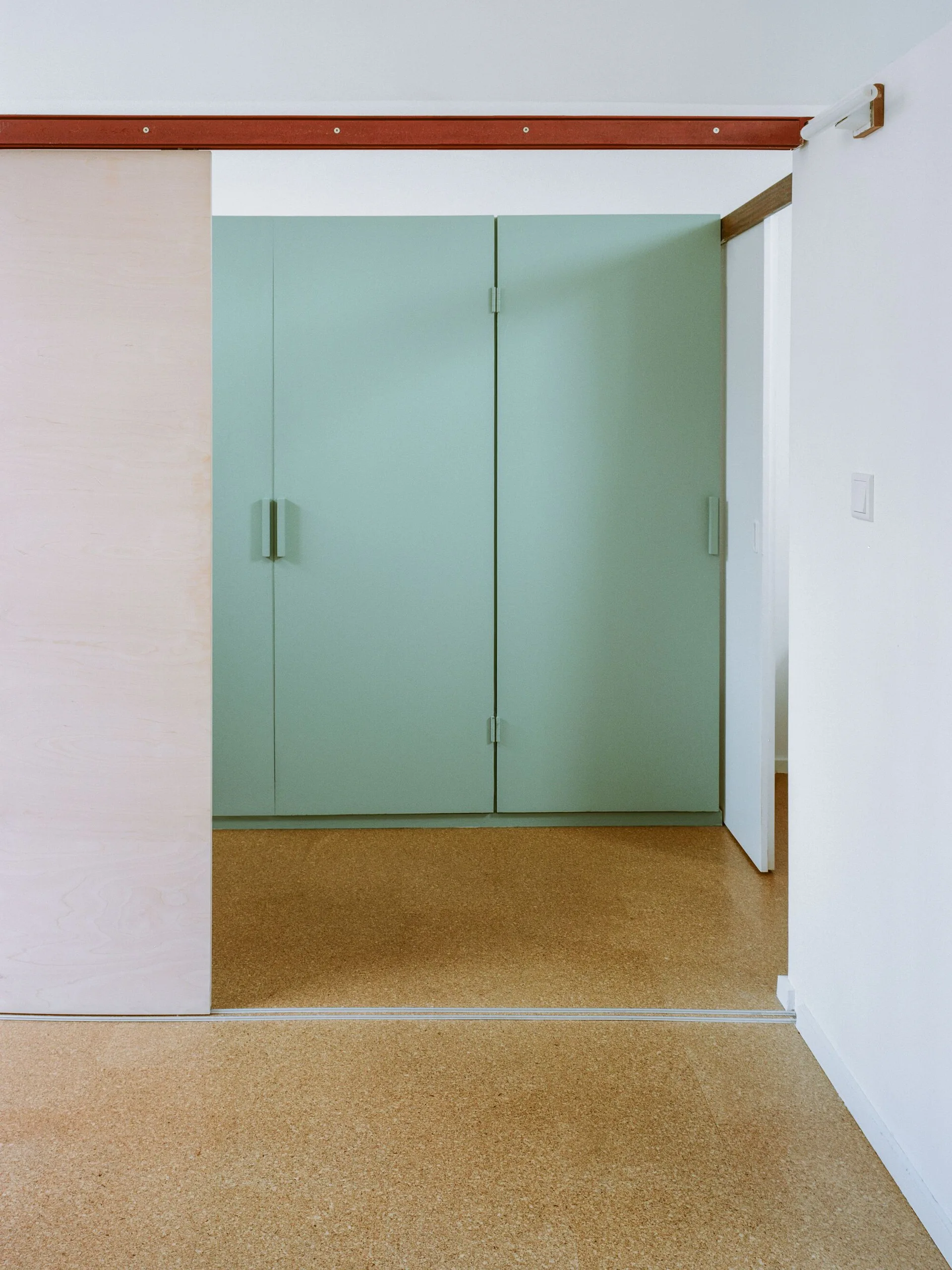 A minimalist hallway features built-in mint green cabinets, a natural plywood sliding door, and textured cork flooring.
