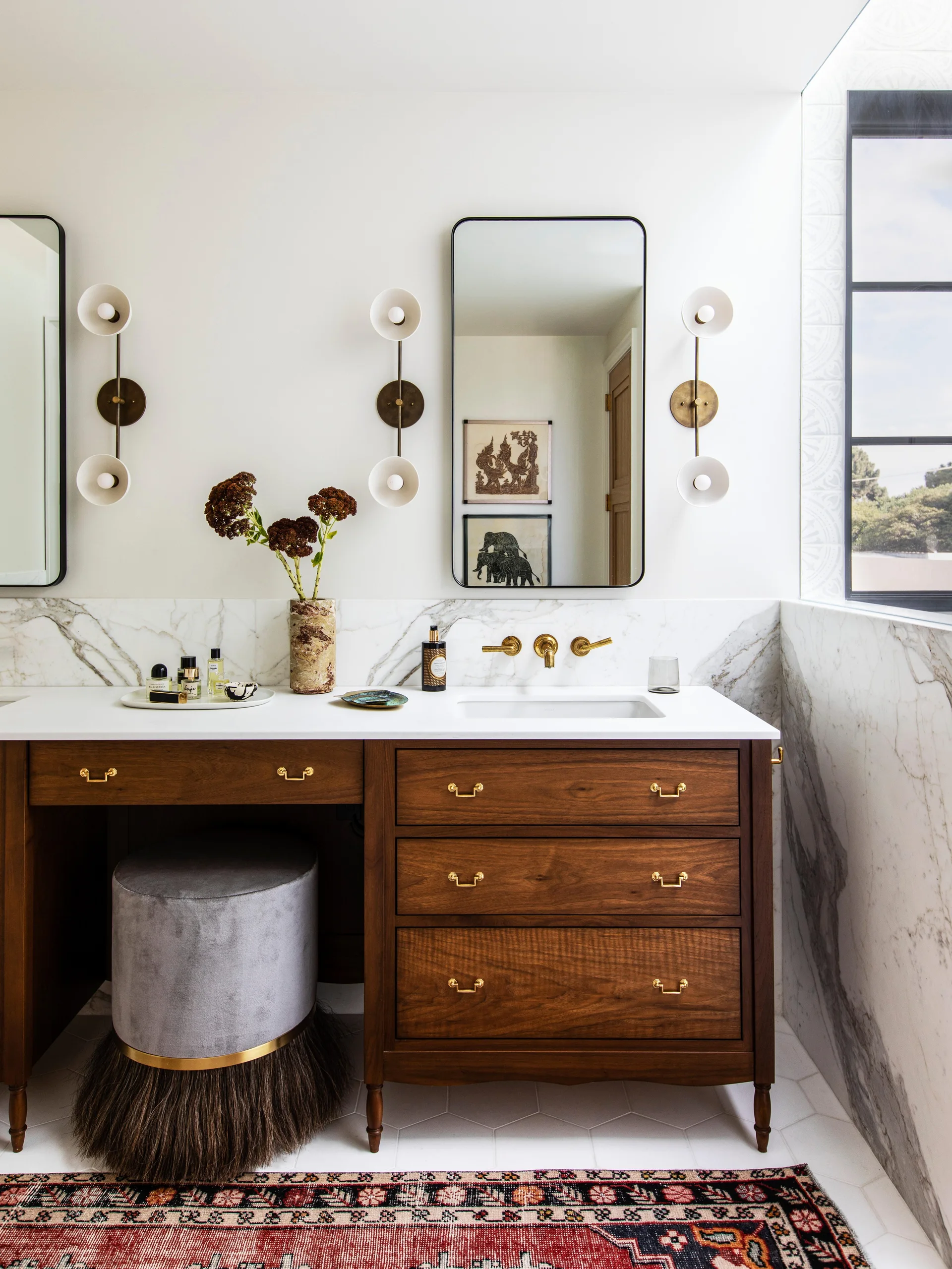 A dark wood bathroom vanity features a marble countertop and backsplash, brass wall-mounted fixtures, and a gray upholstered stool with fringe.
