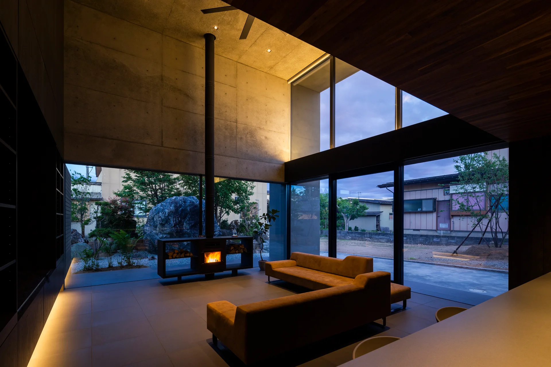A minimalist living room with raw concrete walls, two low-profile brown sofas, a freestanding fireplace with a tall flue, and large glass windows.