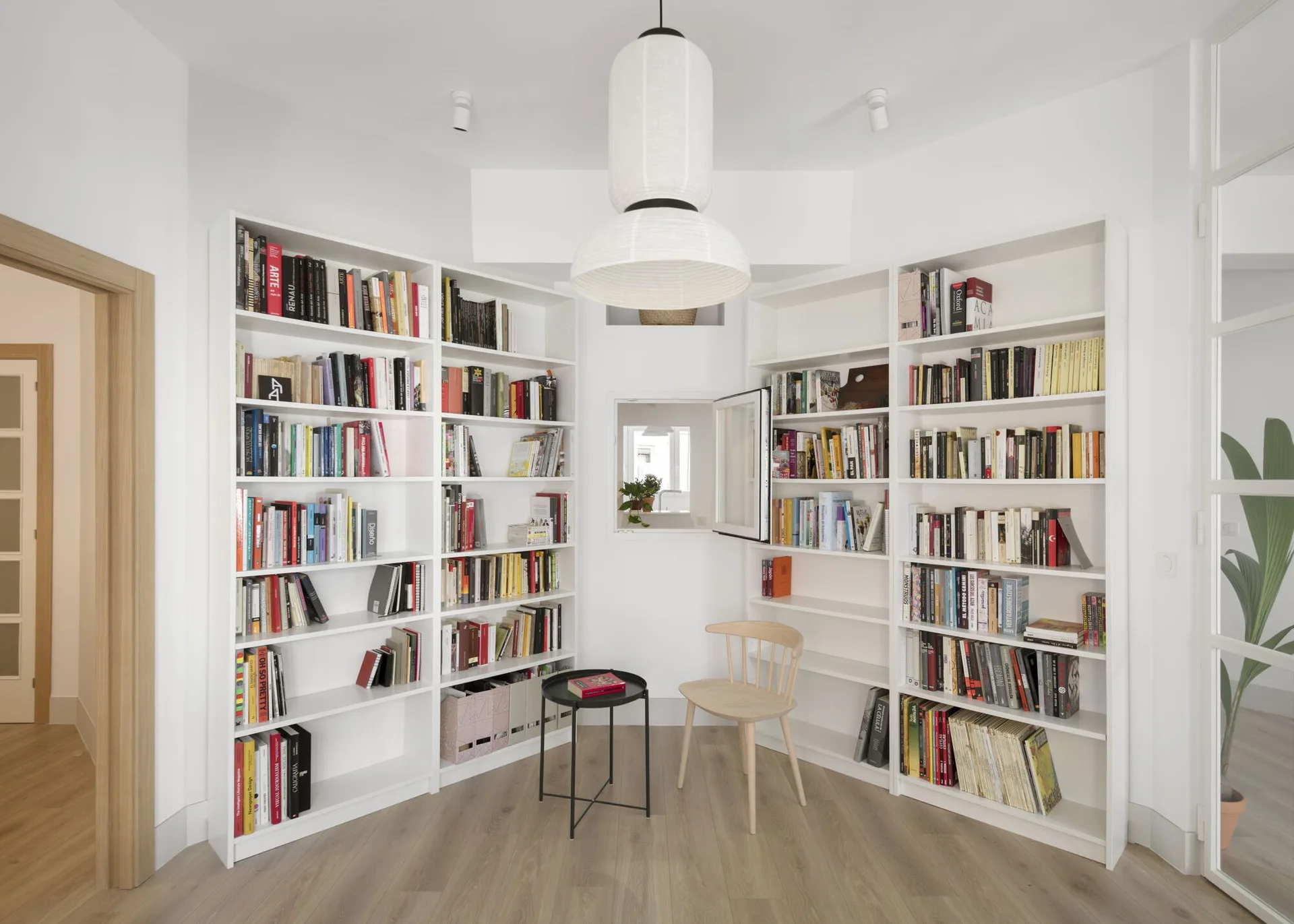 A bright reading corner features white built-in bookshelves surrounding a light wood spindle chair and a black metal side table.