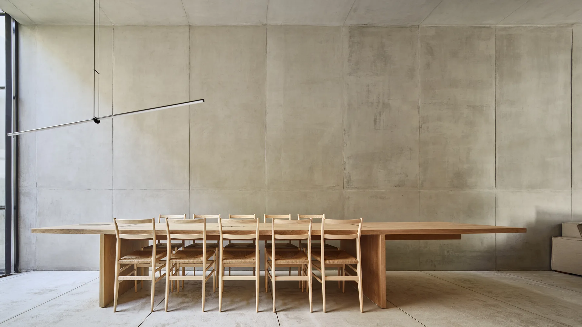 A minimalist dining room features a long light wood dining table with woven-seat chairs set against large concrete wall panels.
