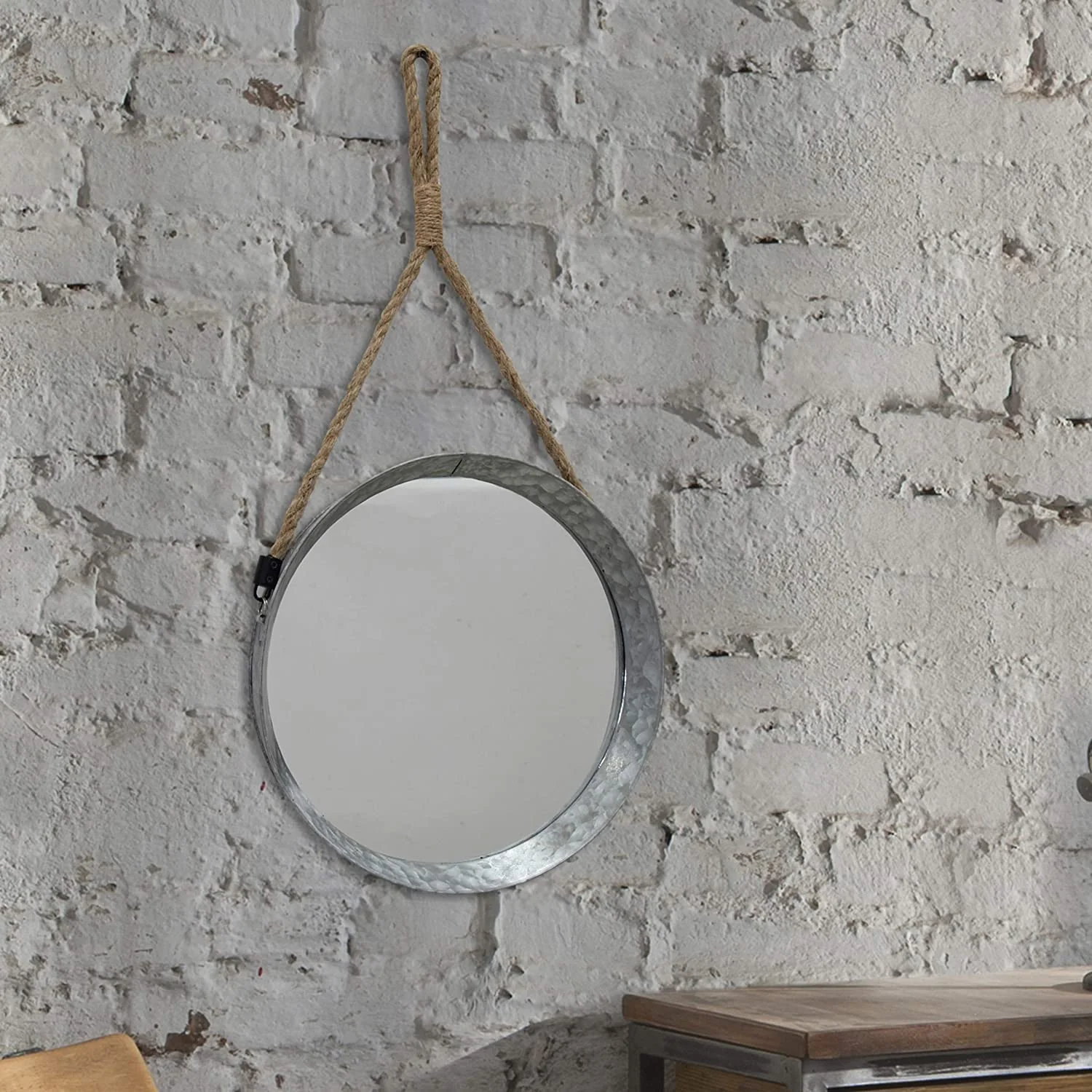 A circular galvanized metal mirror with a natural rope hanger is mounted on a textured white painted brick wall.