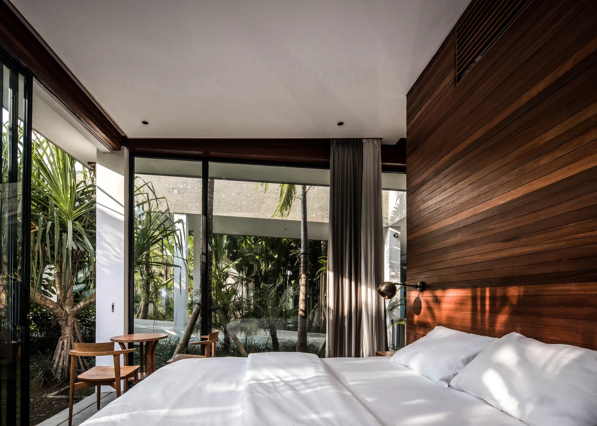 A modern bedroom with a bed with white linens next to a horizontal wood paneled wall and large windows overlooking lush plants.
