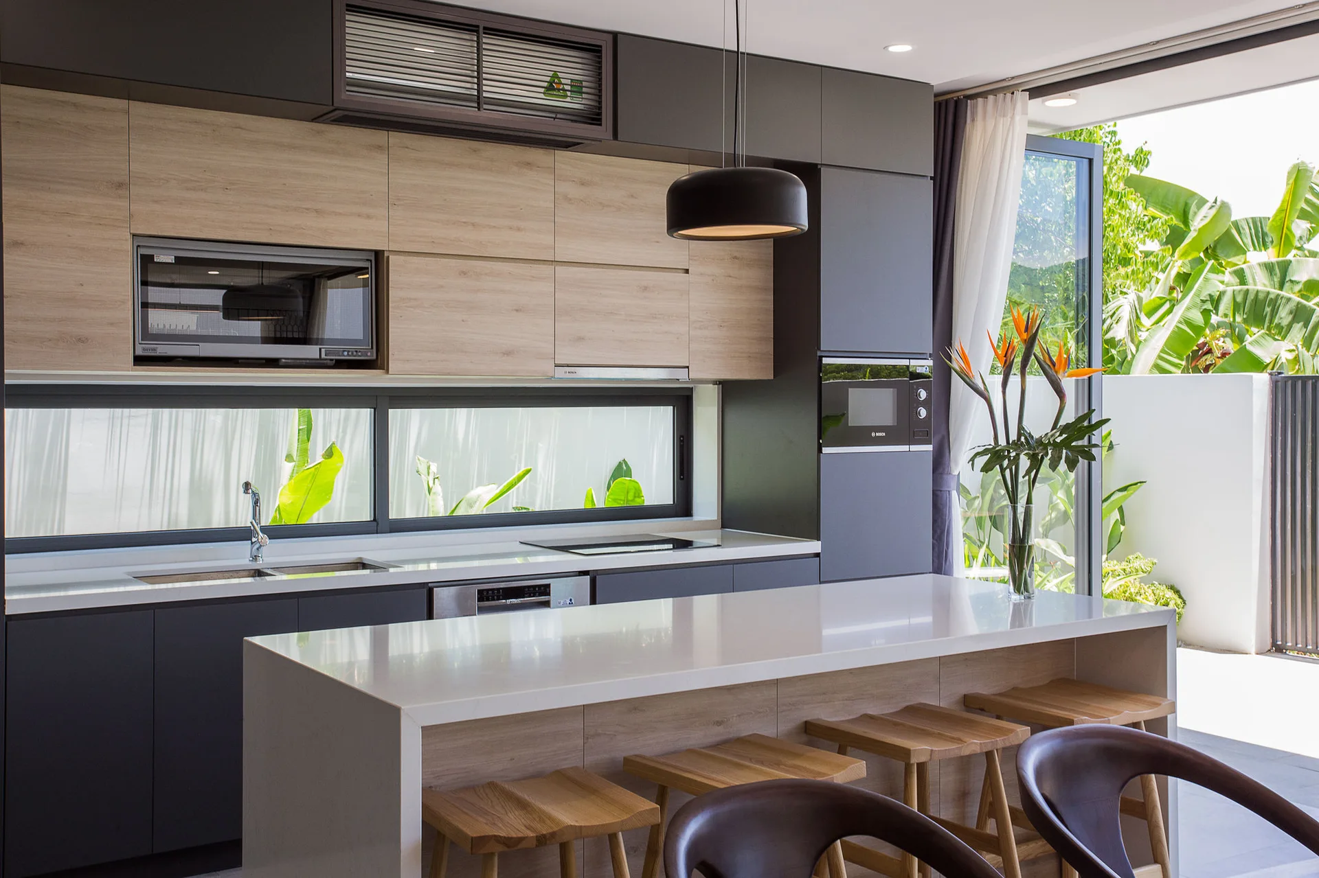 A modern kitchen with matte gray and light wood cabinetry, a large white countertop island with four wooden stools, and a long horizontal window.