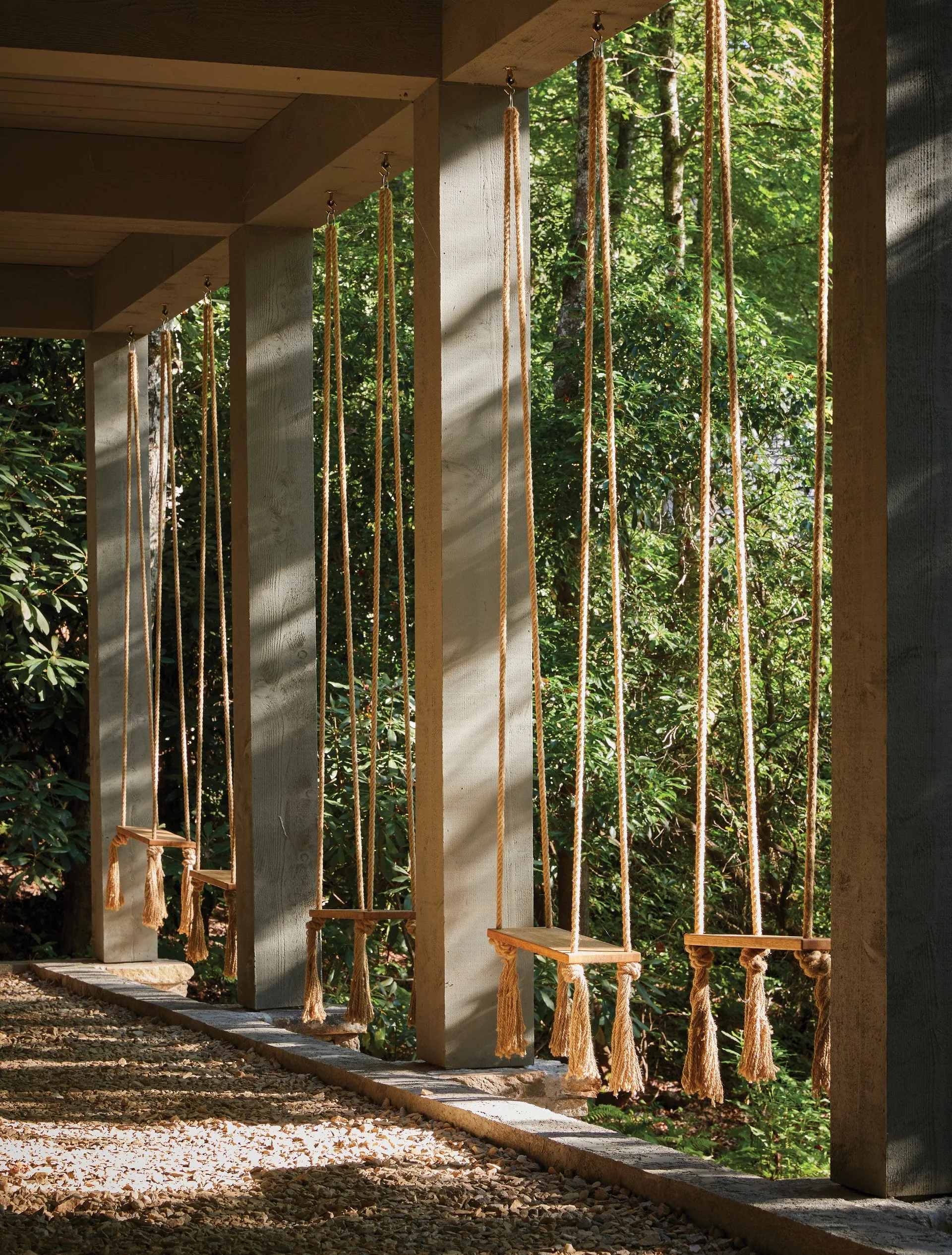A row of wooden rope swings with fringed tassels hangs from a covered grey timber porch overlooking a lush green forest.