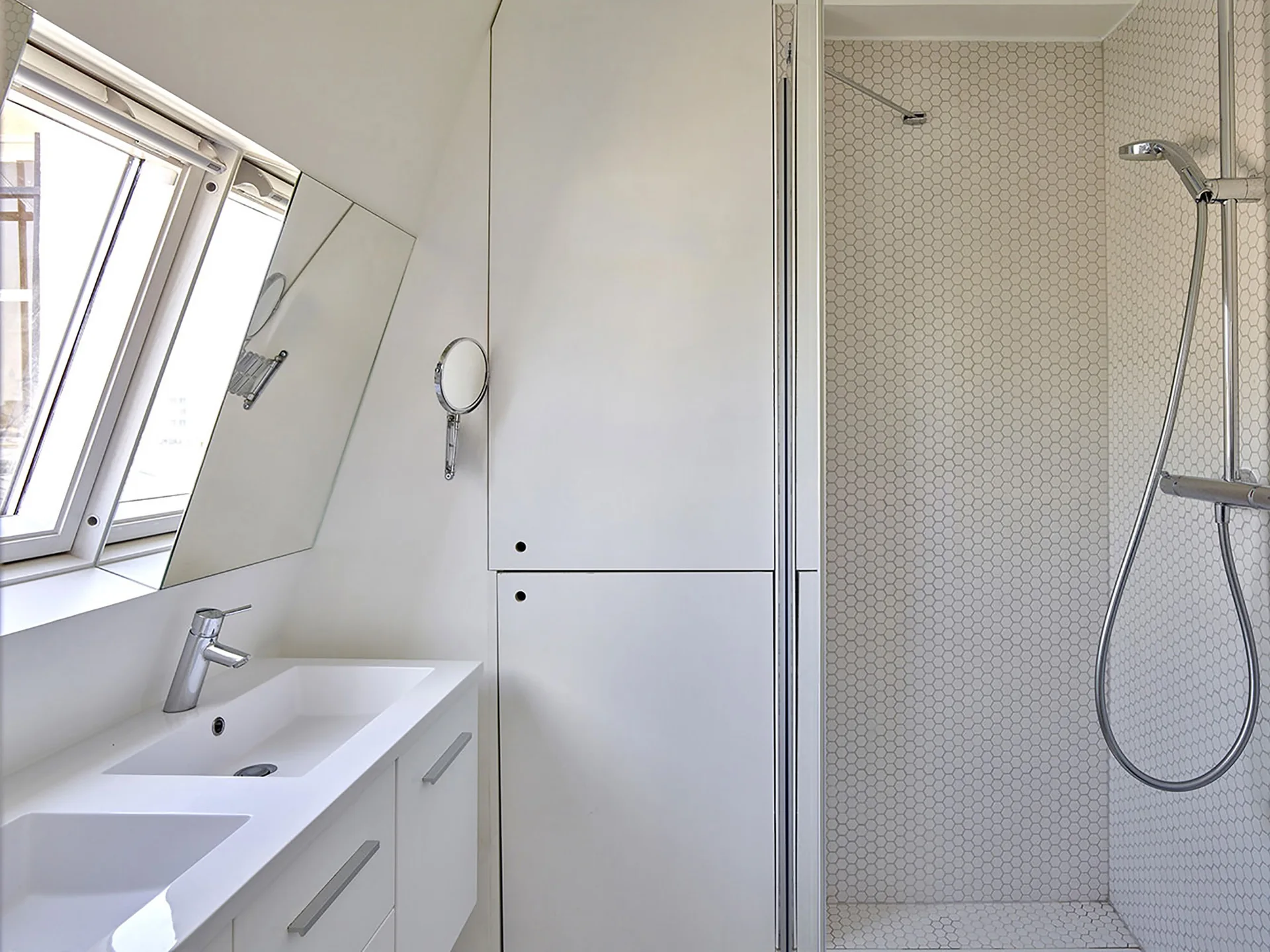 A modern white attic bathroom features a double-sink vanity, a slanted mirror below a sloped window, a tall storage cabinet, and a hex-tiled shower.