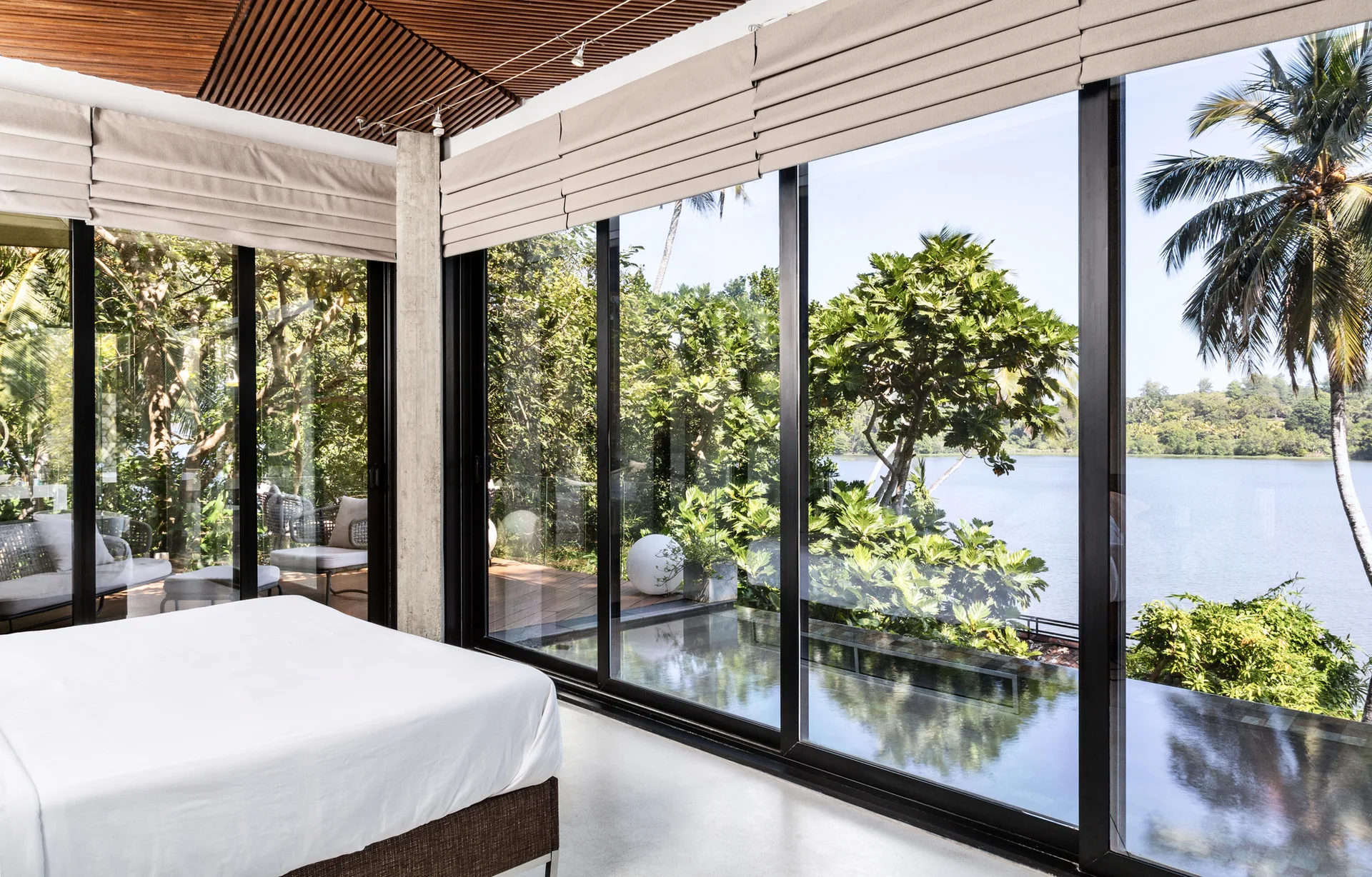 A minimalist bedroom features a bed with white sheets, a slatted wood ceiling, and floor-to-ceiling glass windows overlooking a lake.