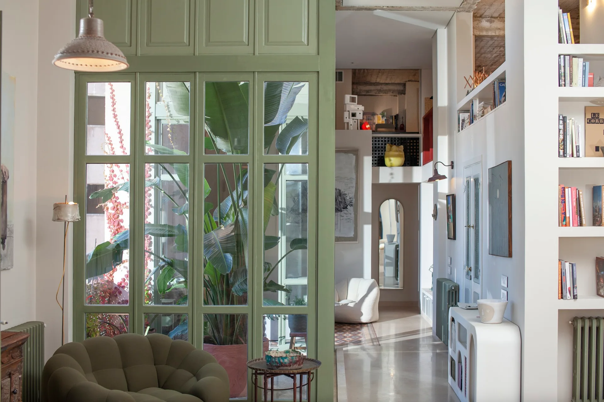 An eclectic living room features a green-framed glass partition wall, a tufted green armchair, and a tall white built-in bookshelf.