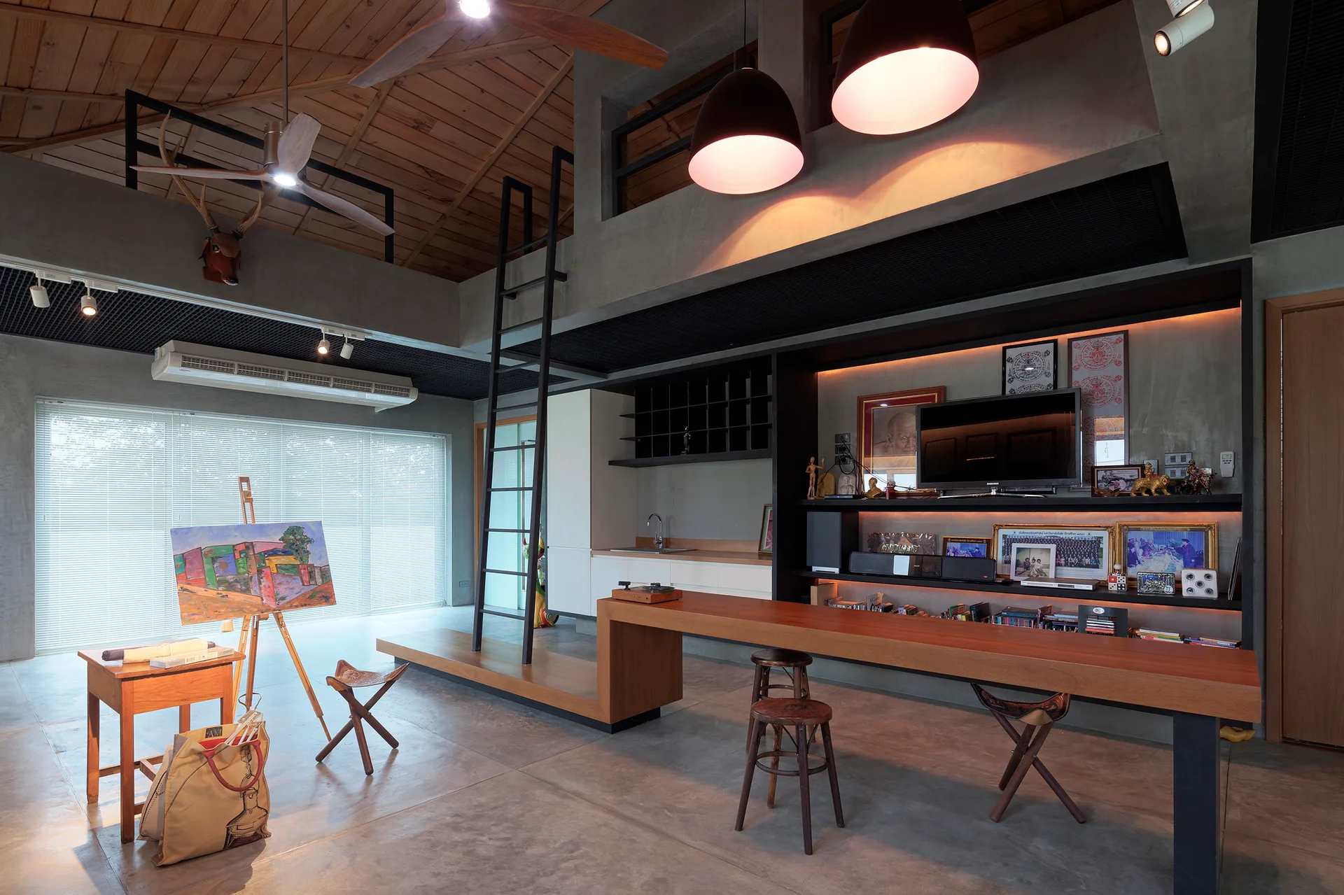 An industrial loft interior features concrete walls and floors, an exposed wood ceiling, a long wooden island with stools, and an art easel.