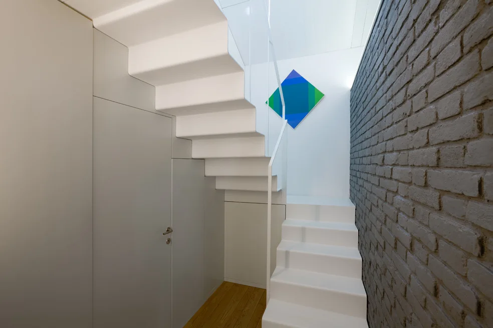 A modern white staircase ascends past a grey painted brick wall and a frameless light grey door built into the wall panels underneath.