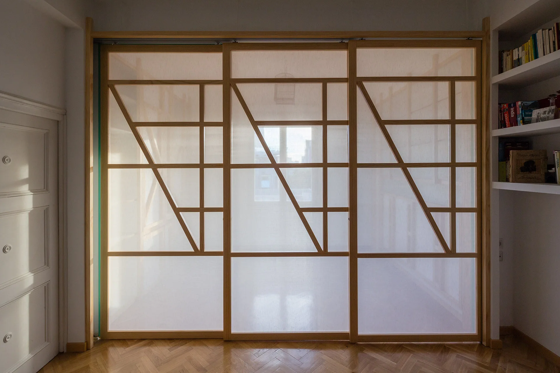 A three-panel shoji-style sliding wooden room divider sits on a herringbone patterned wood floor next to built-in white bookshelves.