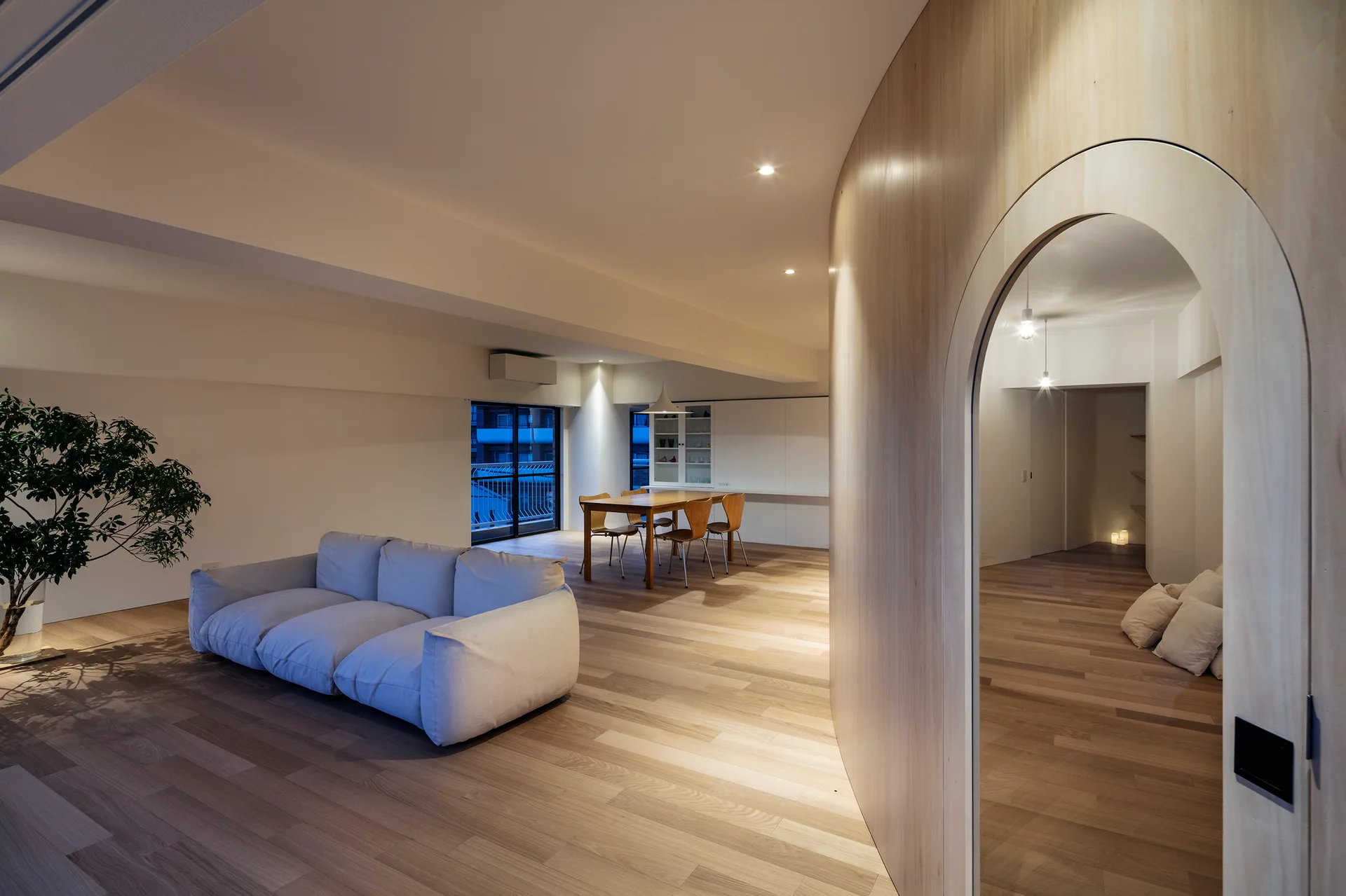 A minimalist living and dining space with light wood floors, a low white modular sofa, a dining table, and a curved wooden wall with an arched doorway.
