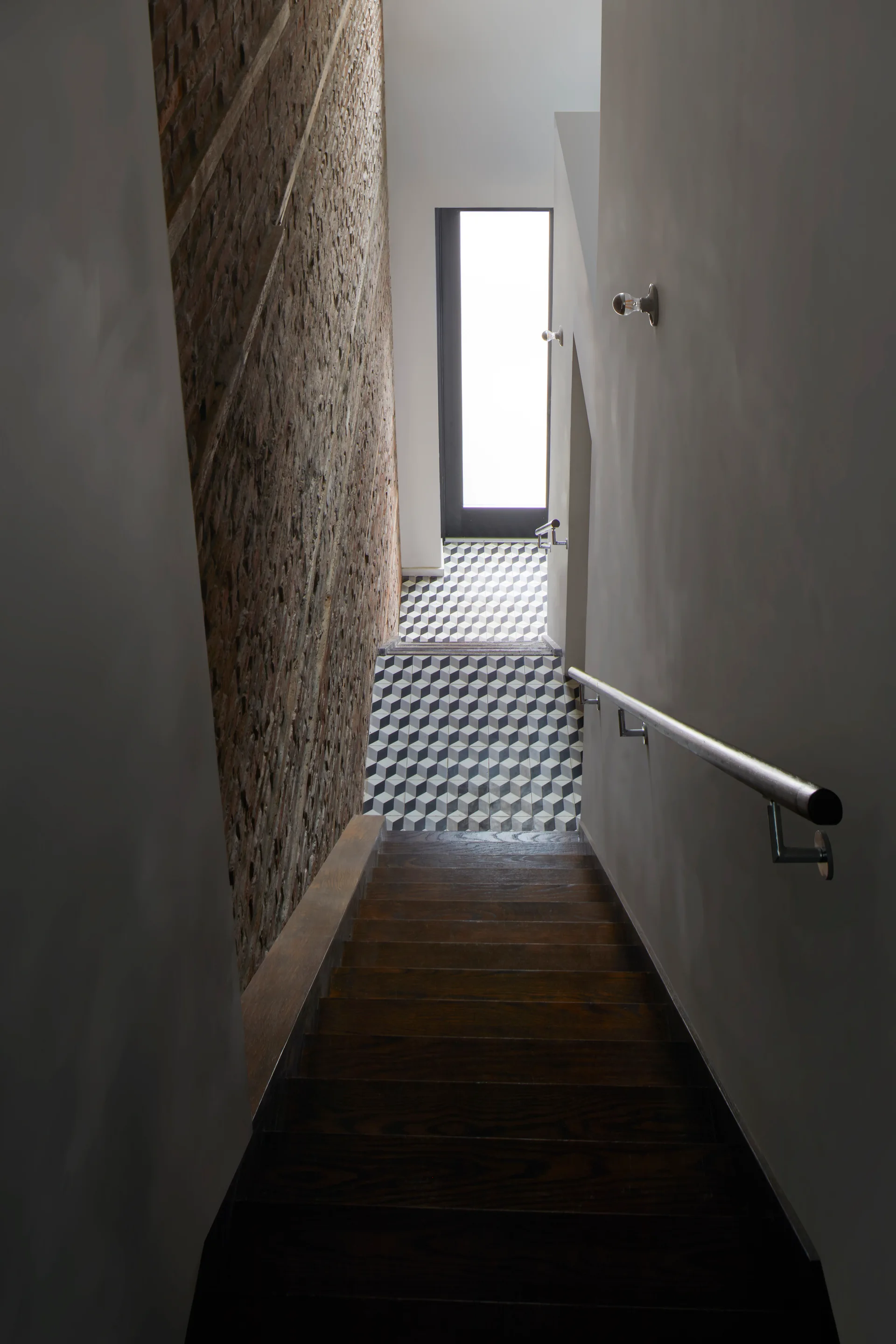 Looking down a wooden staircase bordered by a tall exposed brick wall and a smooth gray wall, leading to a geometric black and white tile landing.
