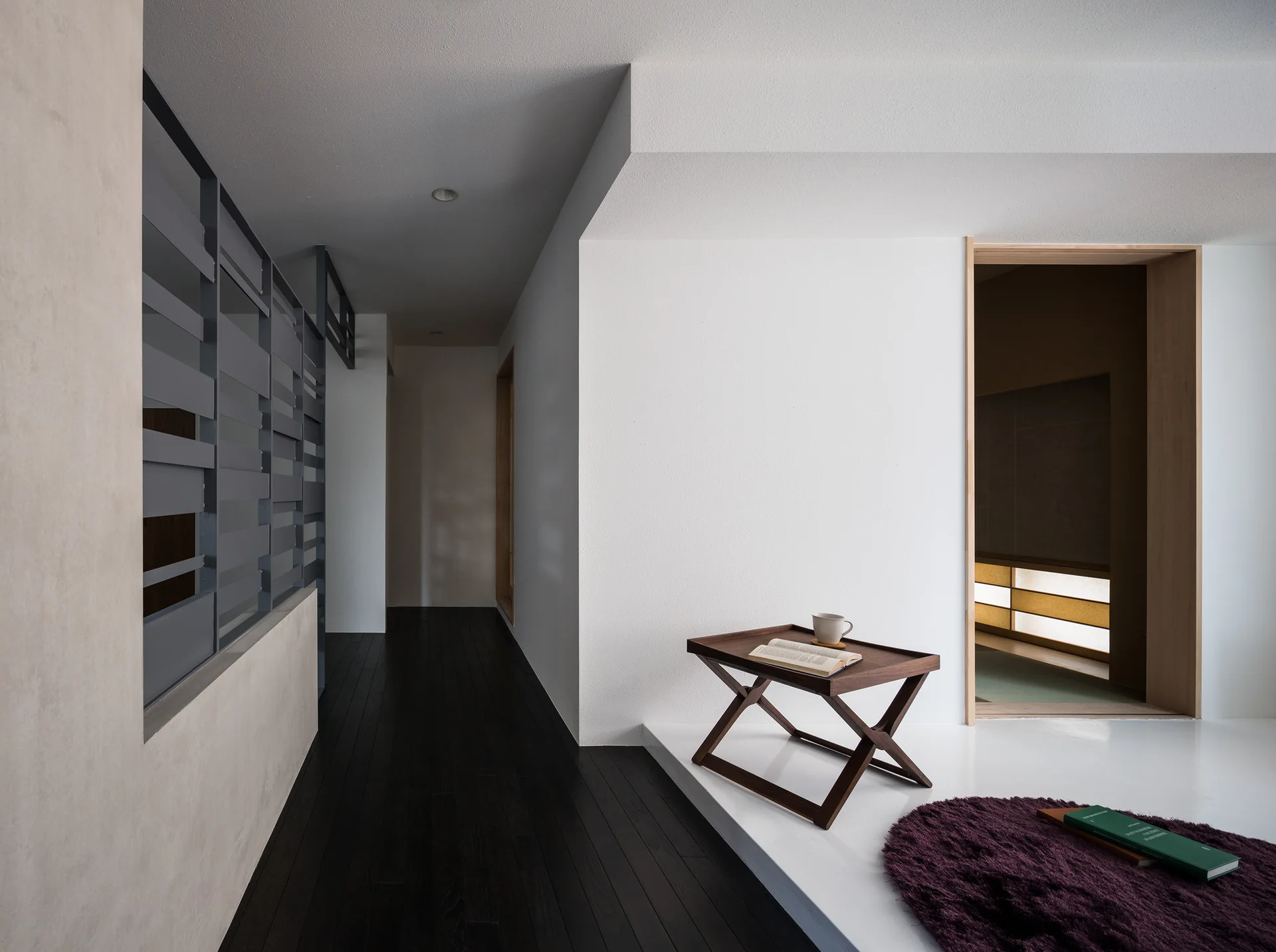 A minimalist hallway features dark wood flooring, a slatted grey room divider, and a raised white platform holding a small folding tray table.