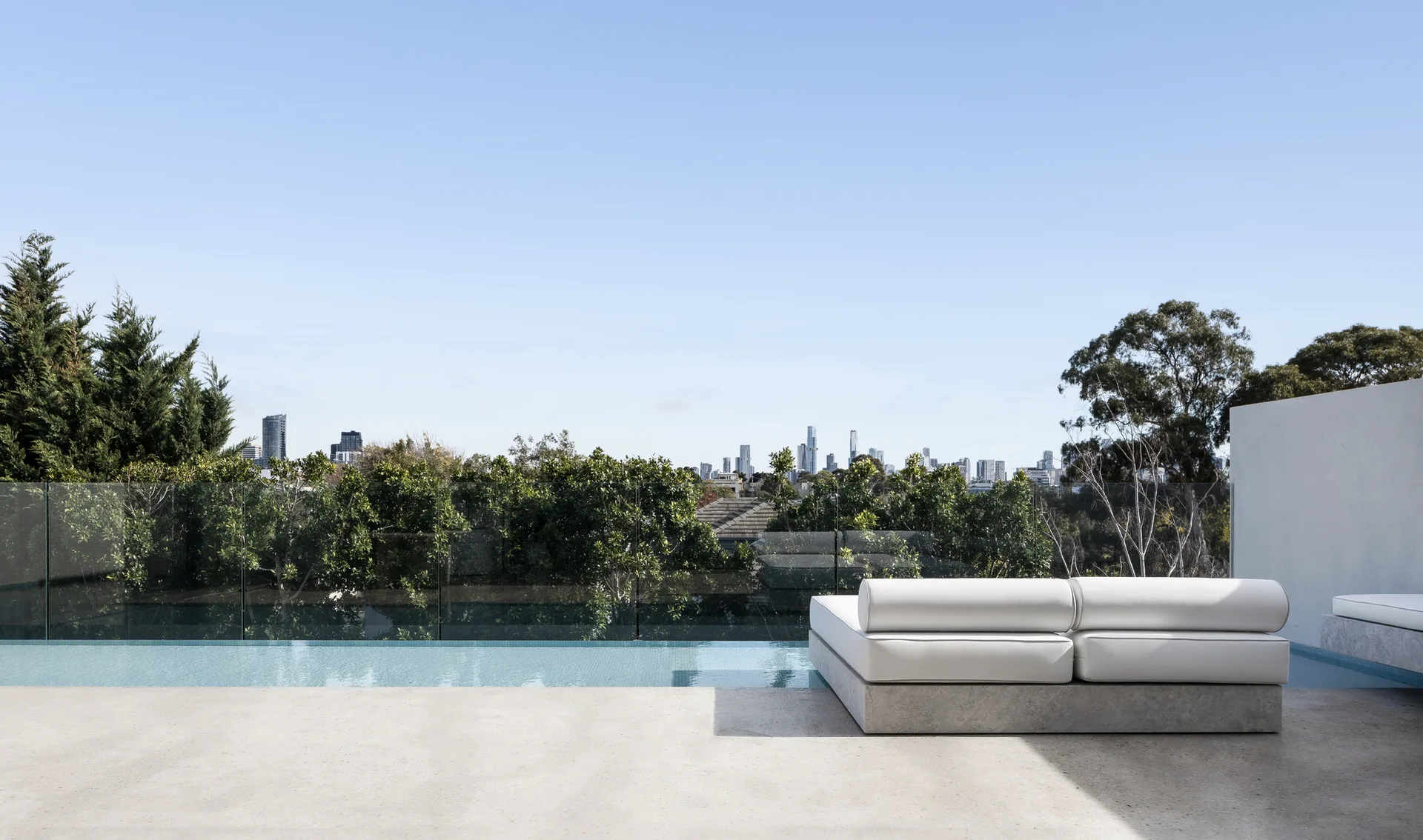 An outdoor concrete terrace features a low white minimalist sofa beside an infinity pool overlooking a green tree line and a distant city skyline.