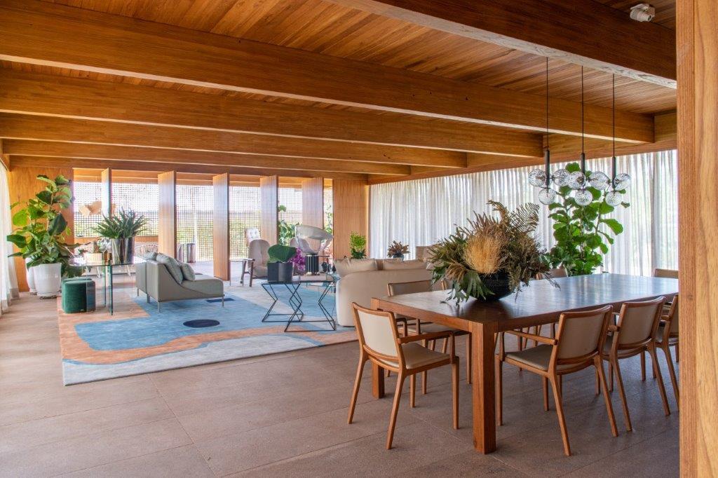 An open-plan dining and living area featuring a wood dining table, matching chairs, an exposed beam ceiling, and a large patterned rug.