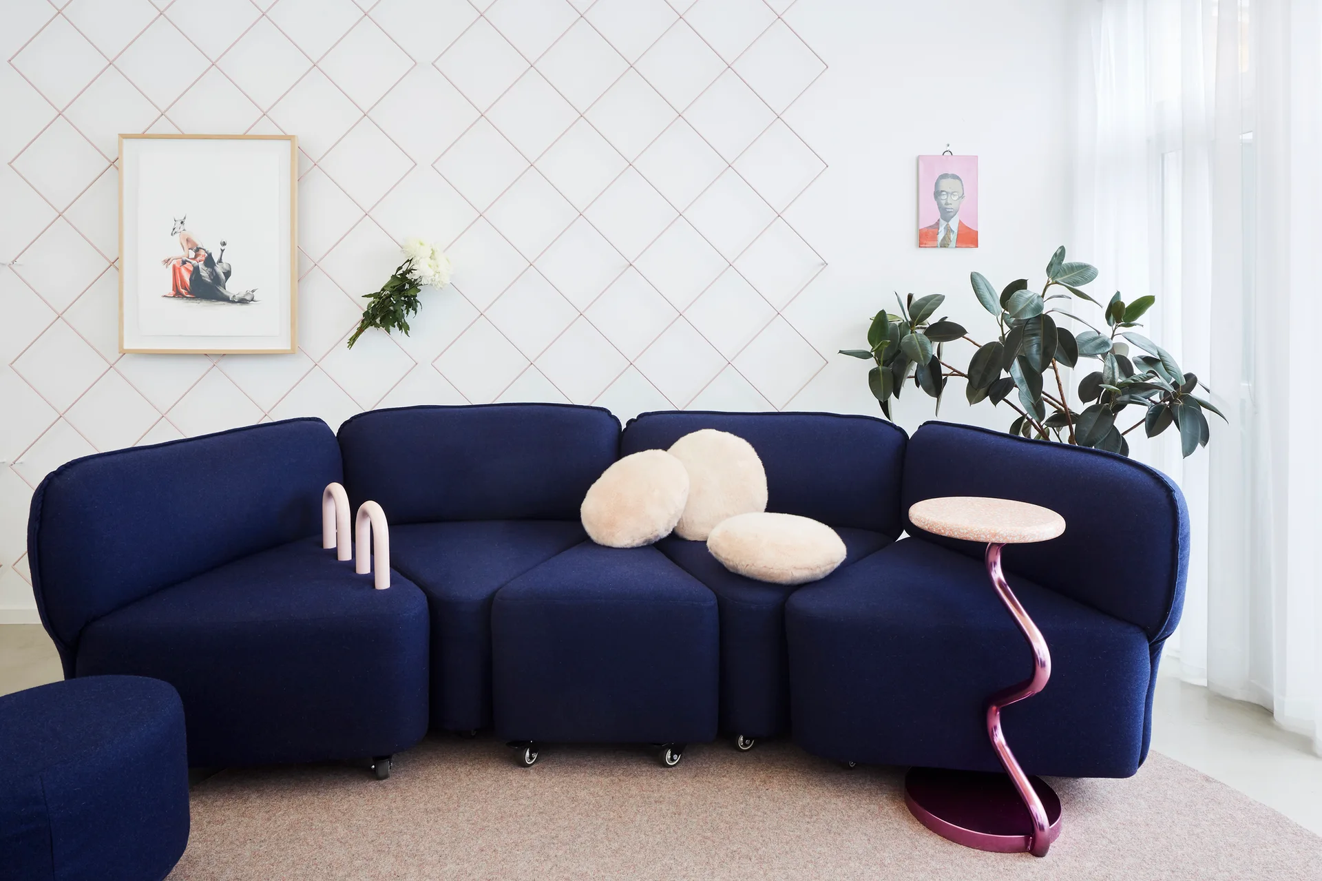 A contemporary living room features a modular navy blue sofa on a textured rug set against a white wall with a pink diamond grid pattern.
