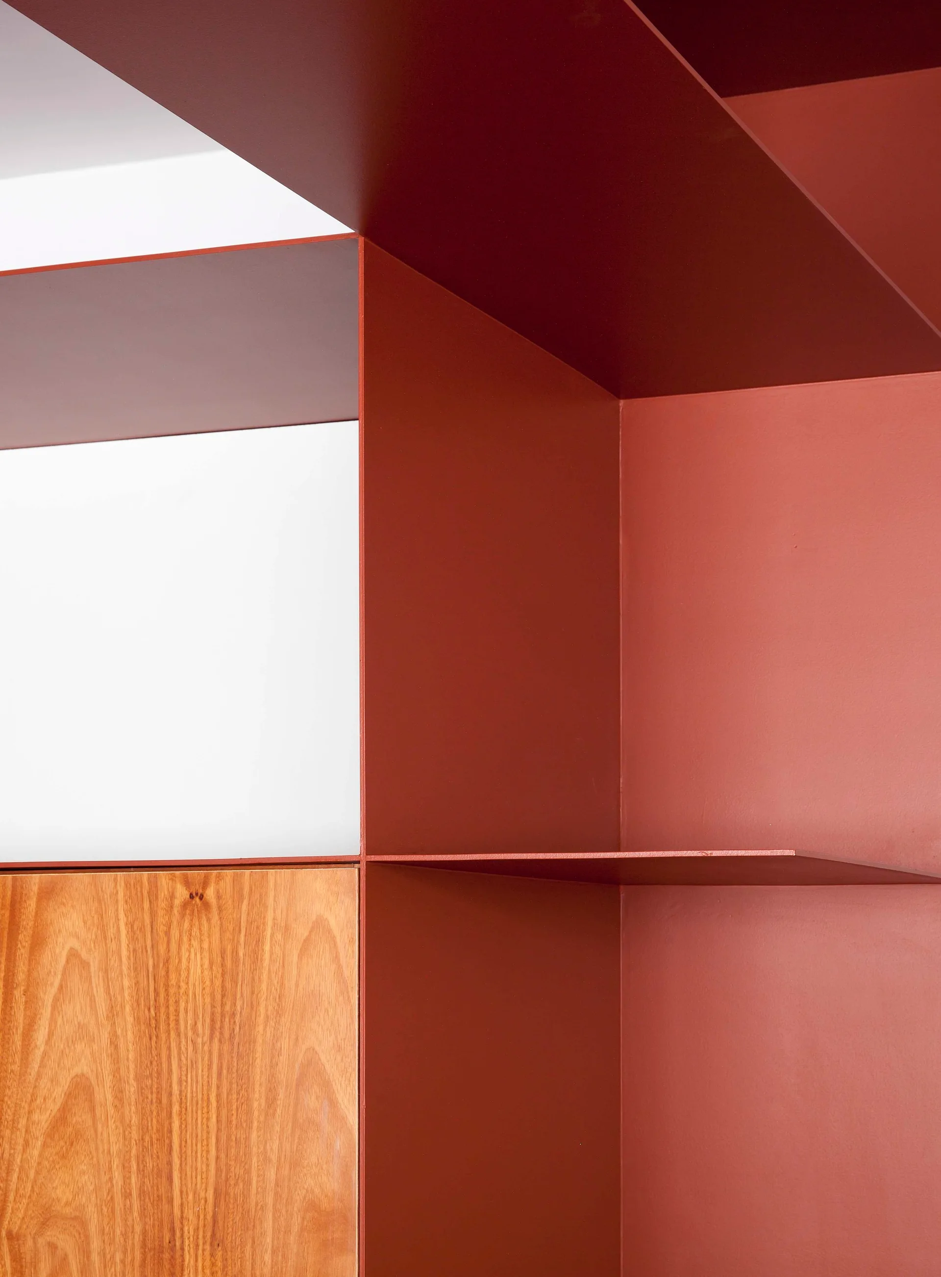 A close-up of geometric built-in shelves in matte terracotta paint next to a warm wood veneer cabinet door and a white panel.