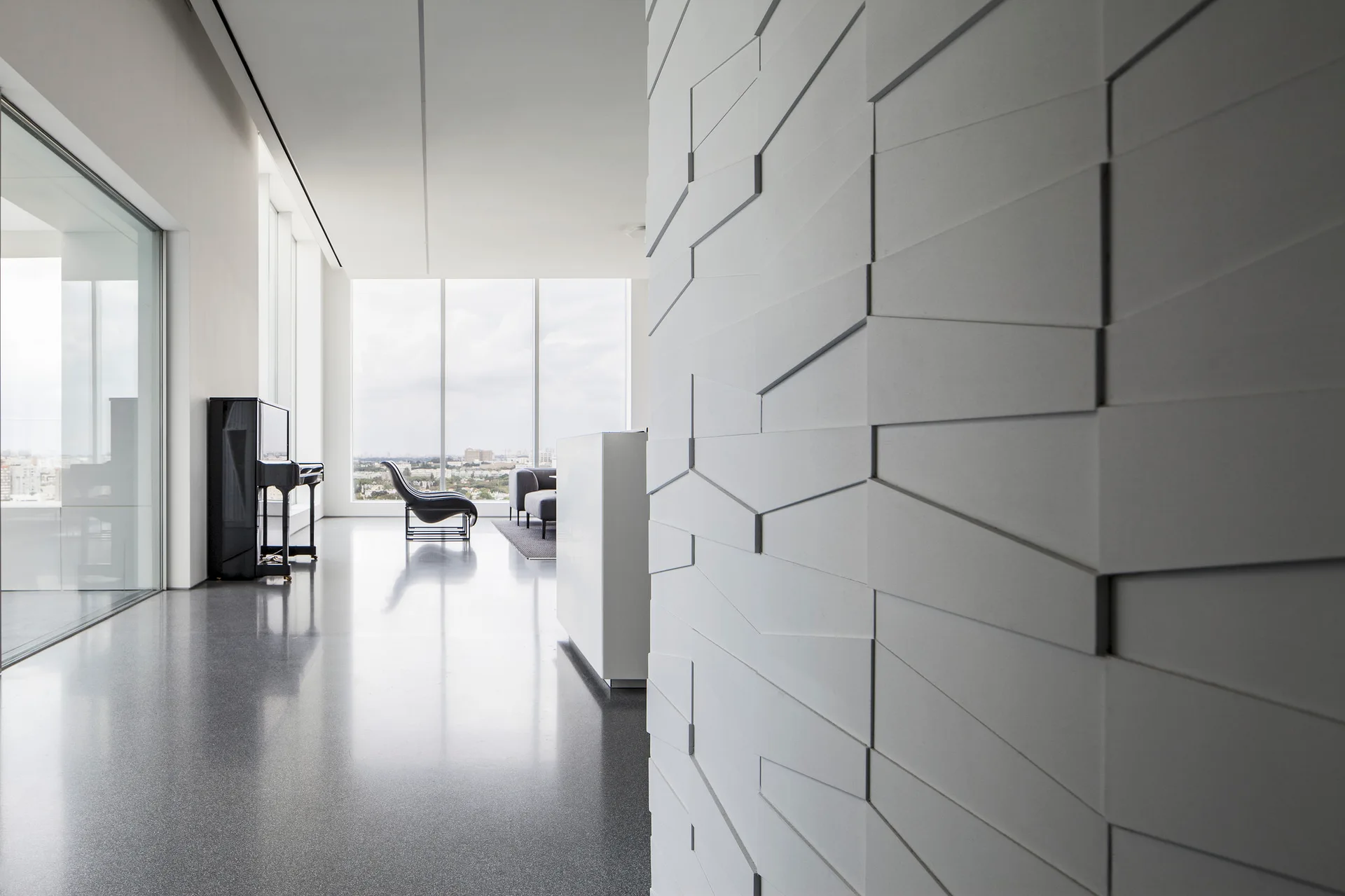 A minimalist modern living room featuring a white geometric accent wall, a polished gray floor, a black upright piano, and large windows.