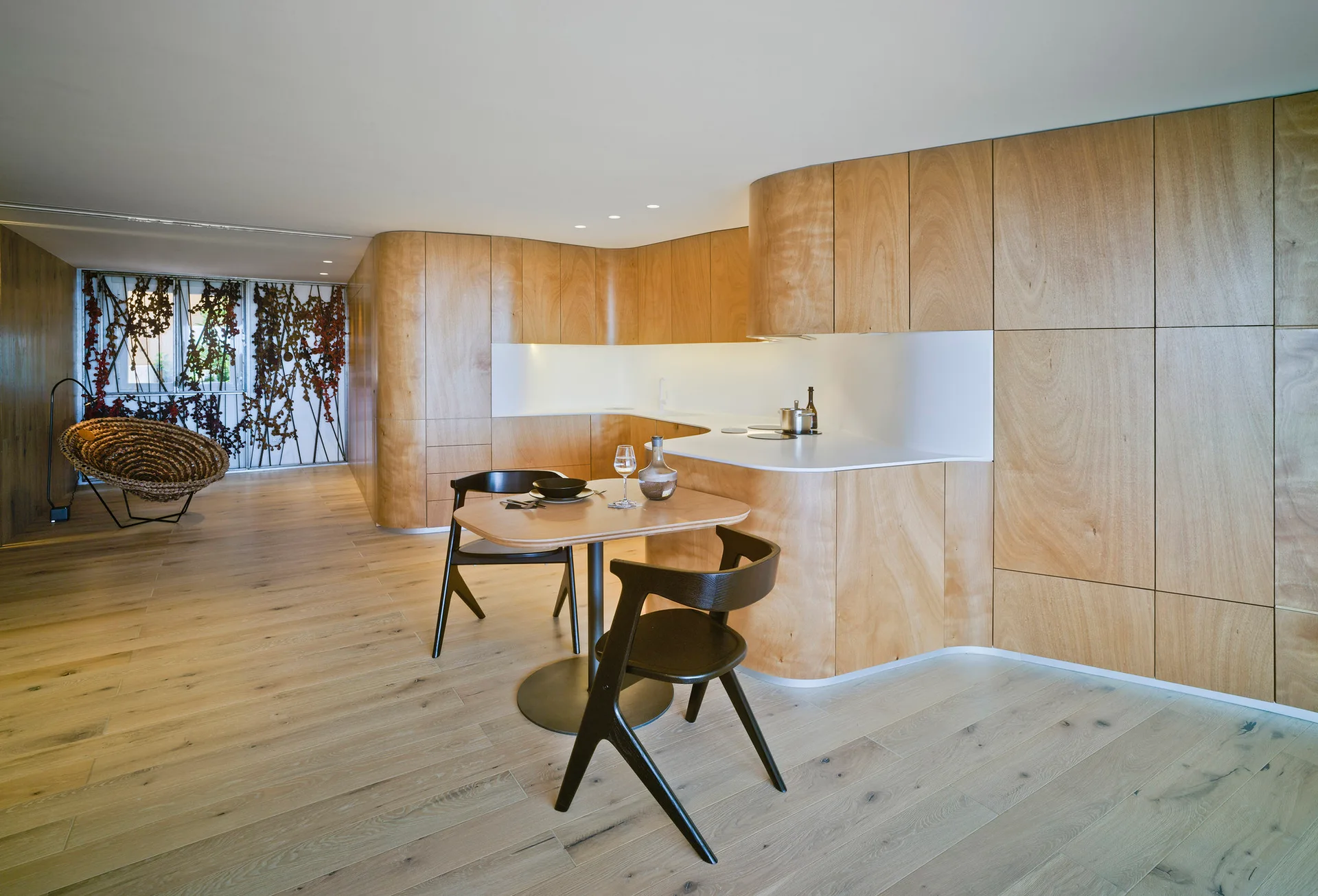 A minimalist kitchen features curved light wood cabinetry paneling, a white countertop niche, and a small dining table with two black chairs.