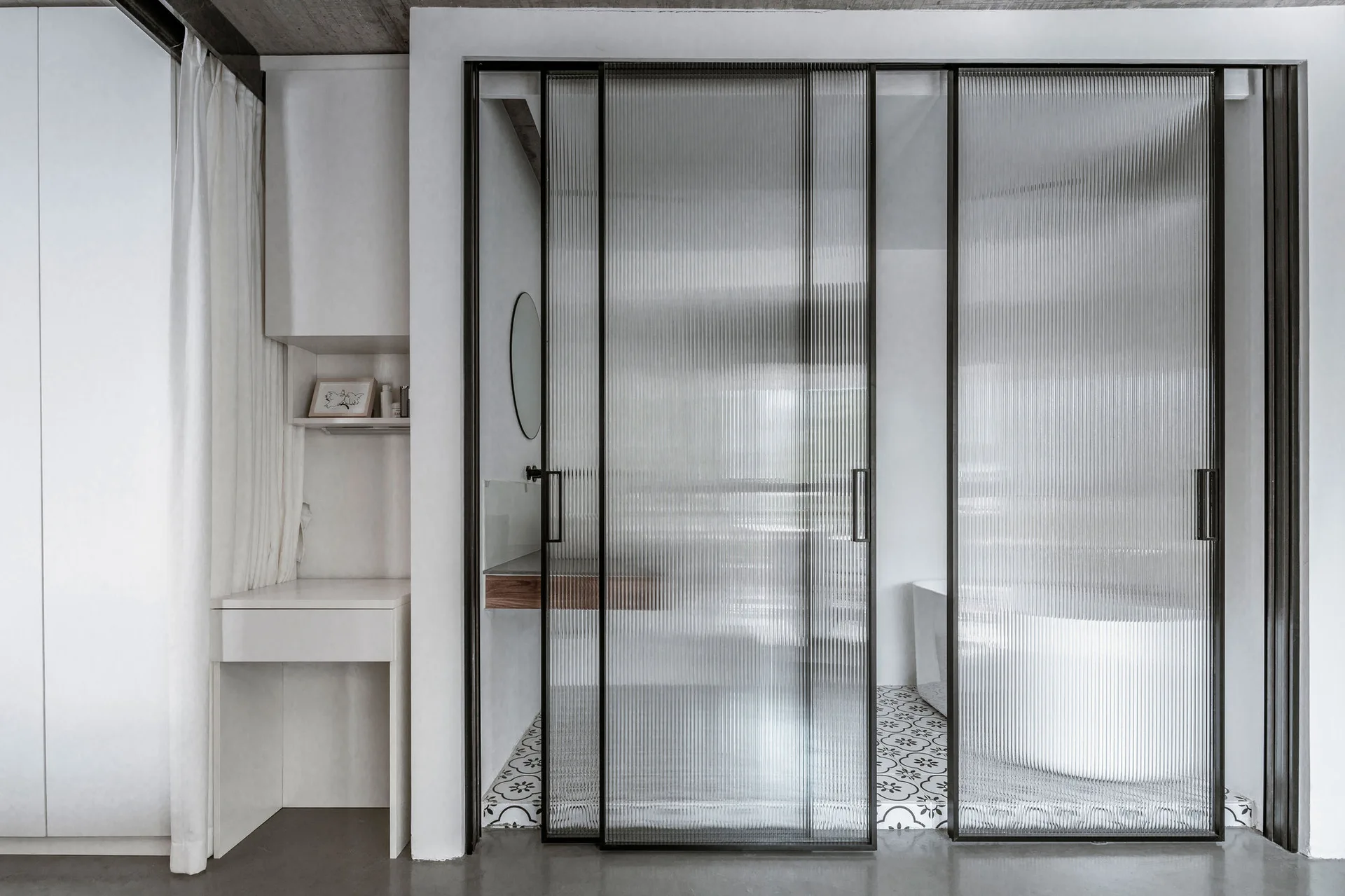 Fluted glass sliding doors in black frames separate a minimalist bathroom with patterned floor tiles from an adjacent white vanity area.