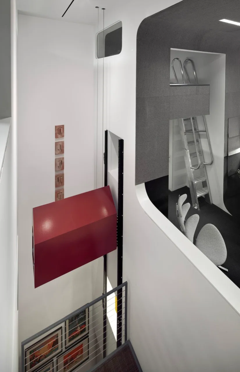 A high-angle view of a modern white stairwell featuring a suspended red rectangular structure and an adjacent mezzanine with a metal ladder.