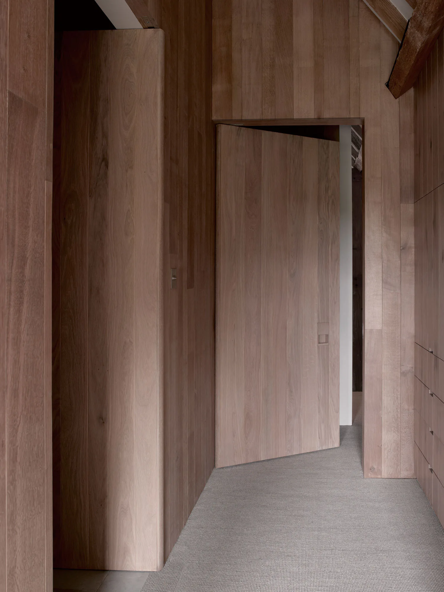 A minimalist hallway lined with vertical wood paneling, featuring a matching flush door and grey woven carpeting.