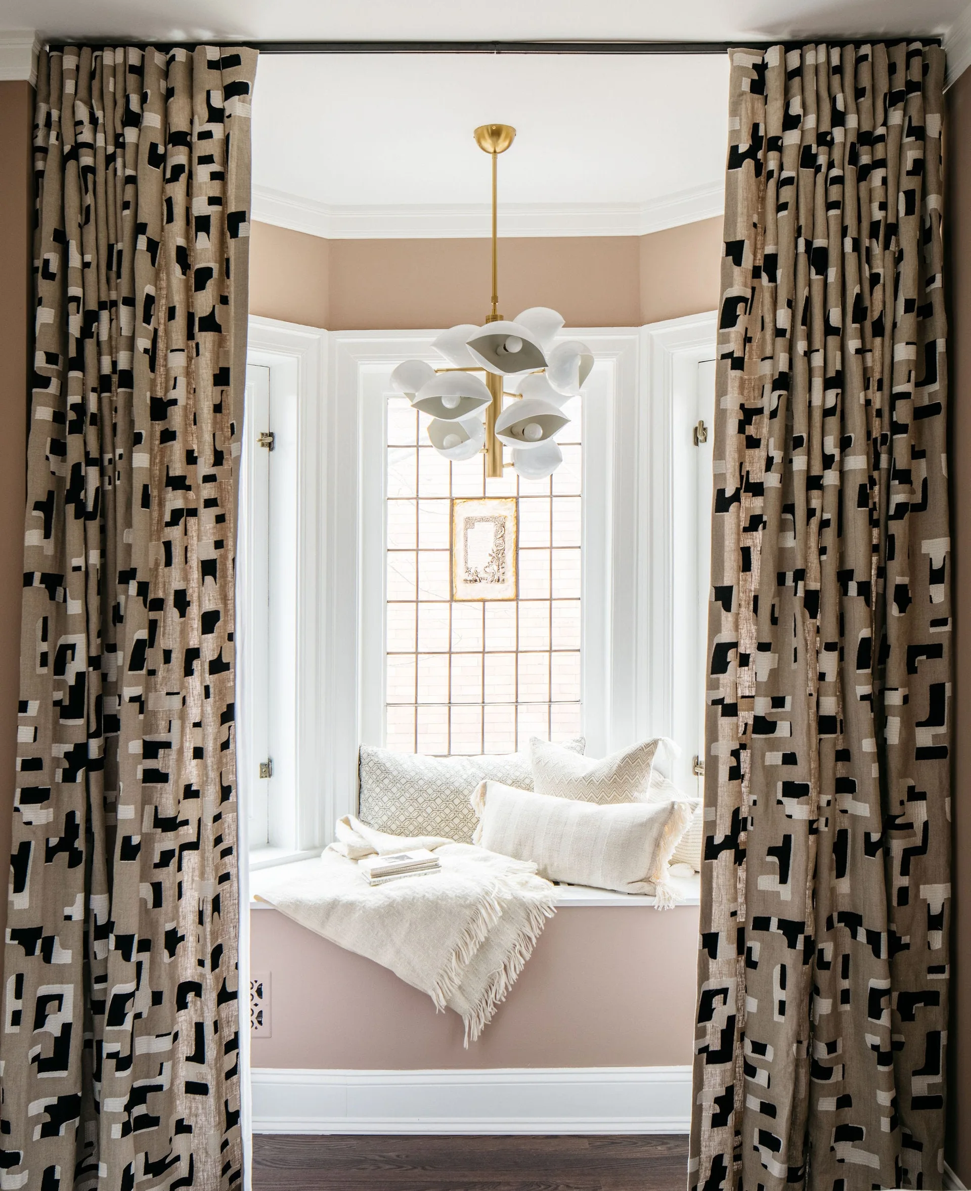 A cozy bay window seat with white cushions and throw pillows is framed by abstract patterned curtains and illuminated by a sculptural white chandelier.